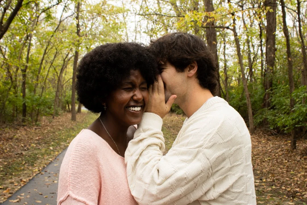 A young man whispering to an older woman, who is smiling and laughing while standing outdoors surrounded by trees.