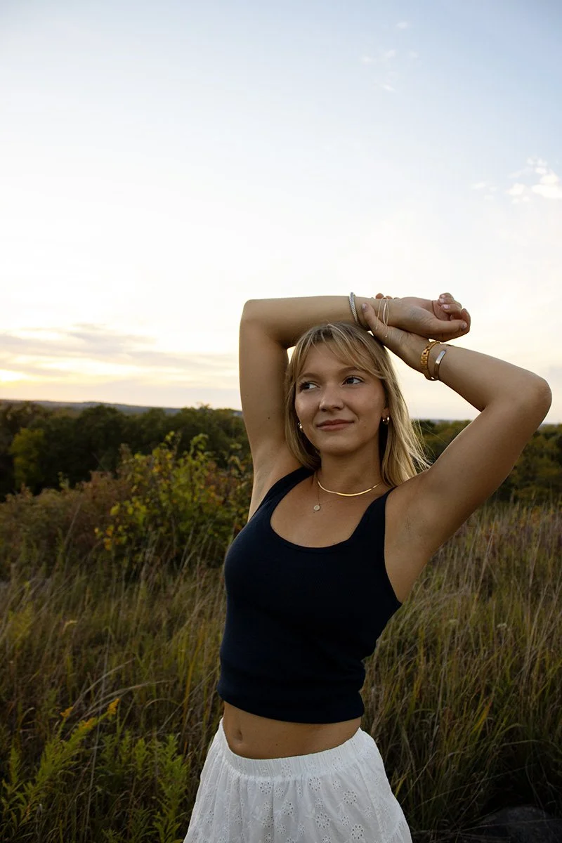 A young woman standing outdoors in a grassy field during sunset, wearing a black tank top and white skirt, with her arms raised above her head and smiling softly.