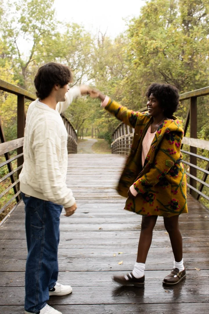 A young man and woman is on a wooden bridge in a park, playing a game with their hands, smiling, surrounded by trees with fall foliage.