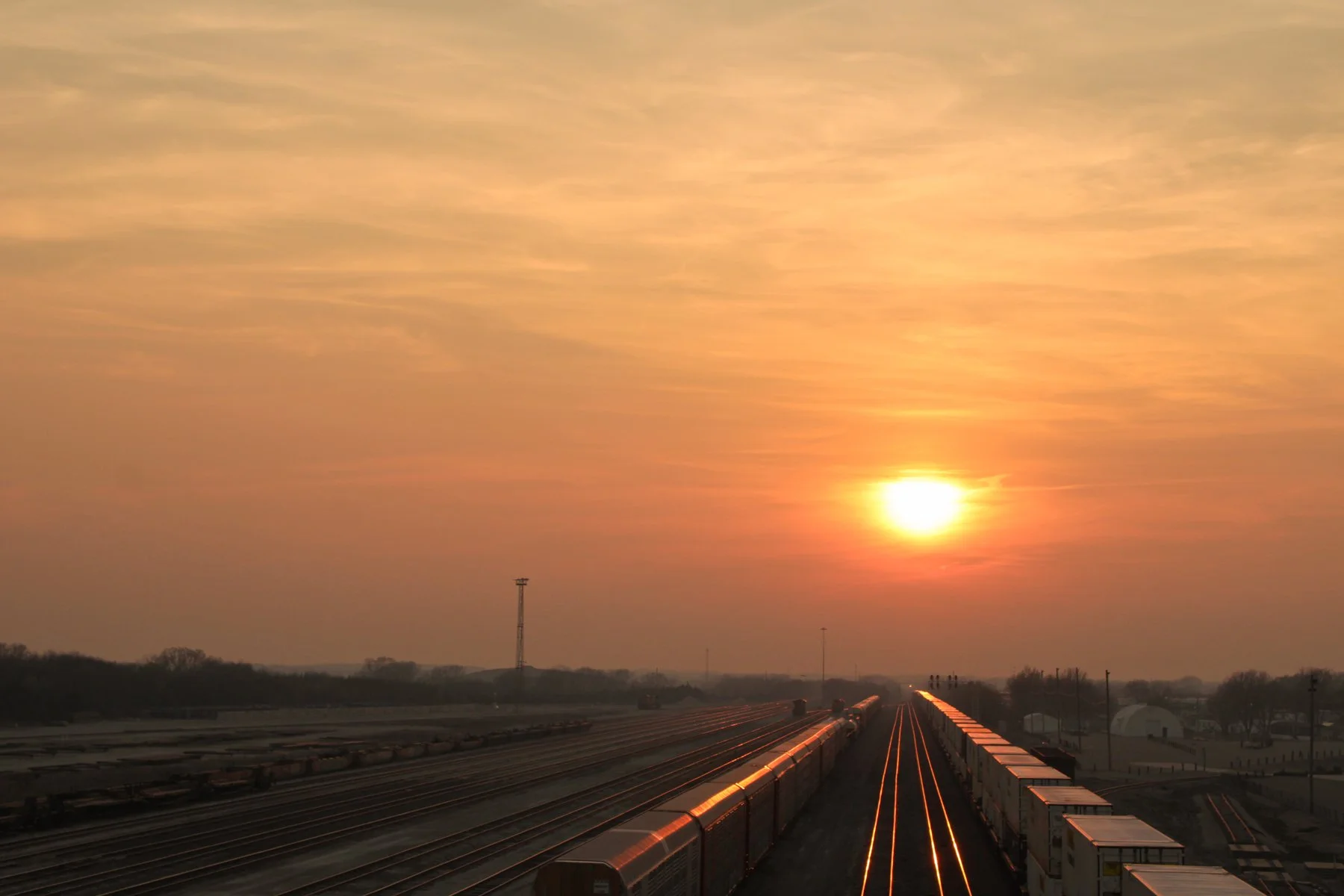 Sunset over a train yard with multiple train tracks and train cars, some of which are illuminated, with a few people visible in the distance and a partly cloudy sky.