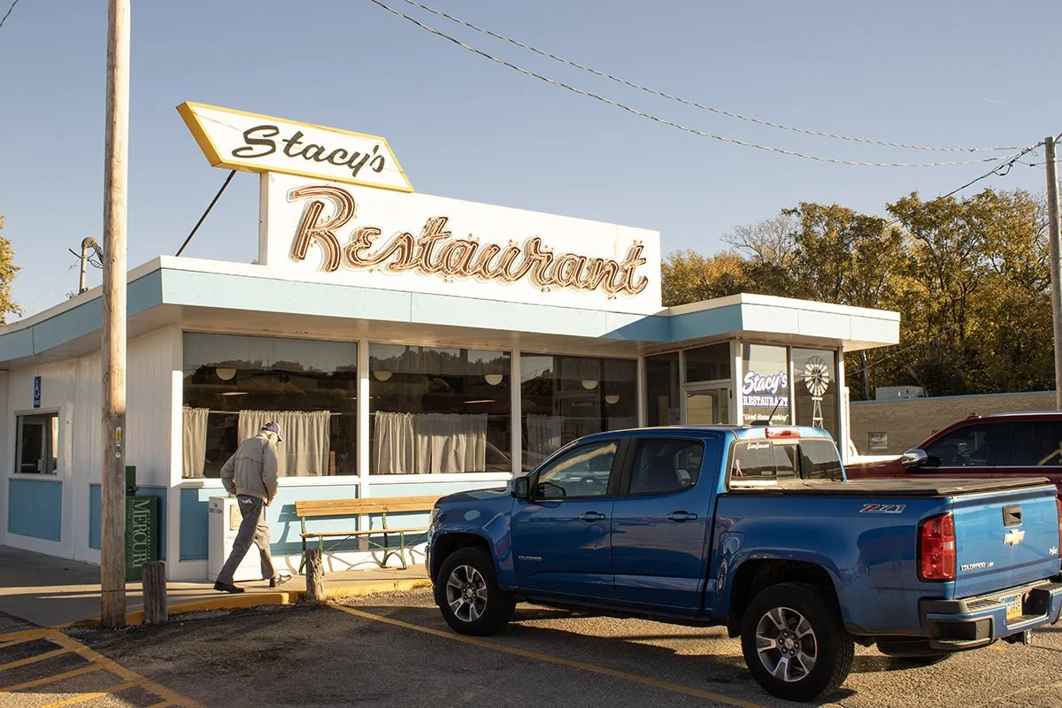 A roadside restaurant called Stacy's Restaurant with a vintage sign. A man walks past the restaurant, and a blue Chevrolet Colorado Z71 pickup truck is parked in front. The building has large windows with curtains, and there are trees and utility pol