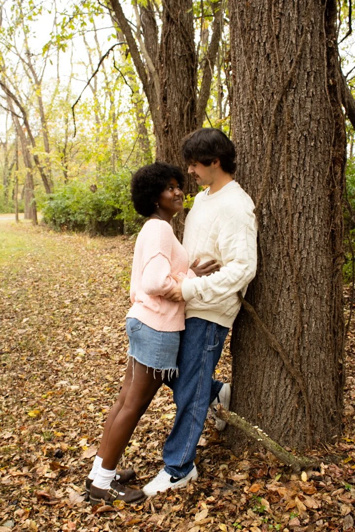 A young couple standing close together in a park, leaning against a large tree, surrounded by fallen leaves, with the woman holding the man's arm and both smiling at each other.