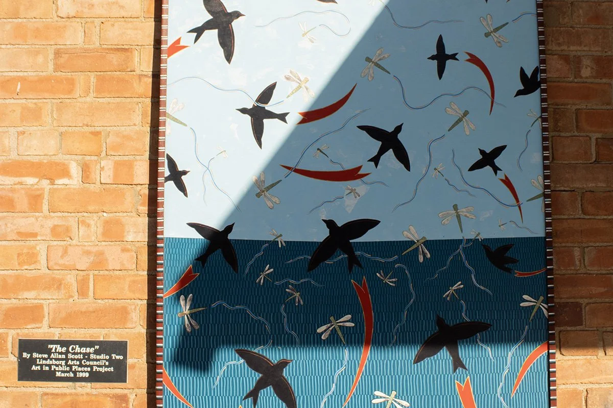 Mural titled 'The Chase' depicting black birds and dragonflies flying over ocean waves, with a light blue sky in the background, mounted on a brick wall.