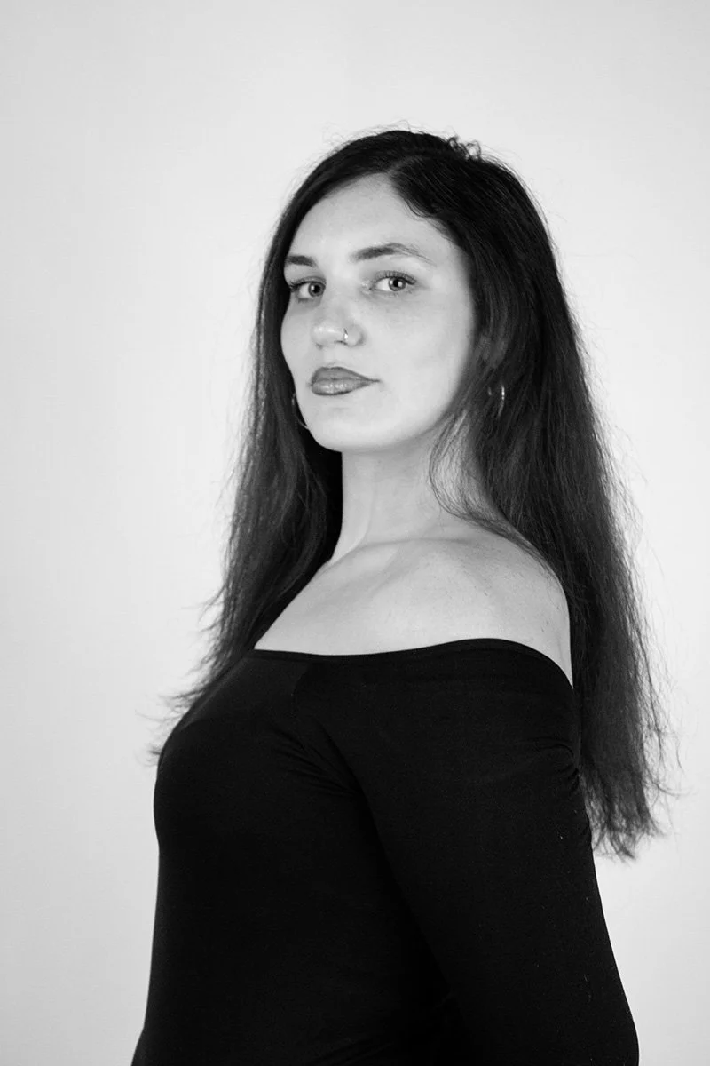 Black and white portrait of a woman with long dark hair, wearing a black off-the-shoulder top, standing against a plain light background.