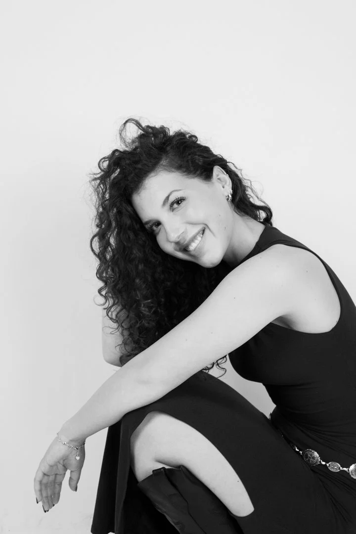 A woman with curly hair wearing a sleeveless black dress, smiling and sitting on the floor with one knee up, in a studio with a plain white background.