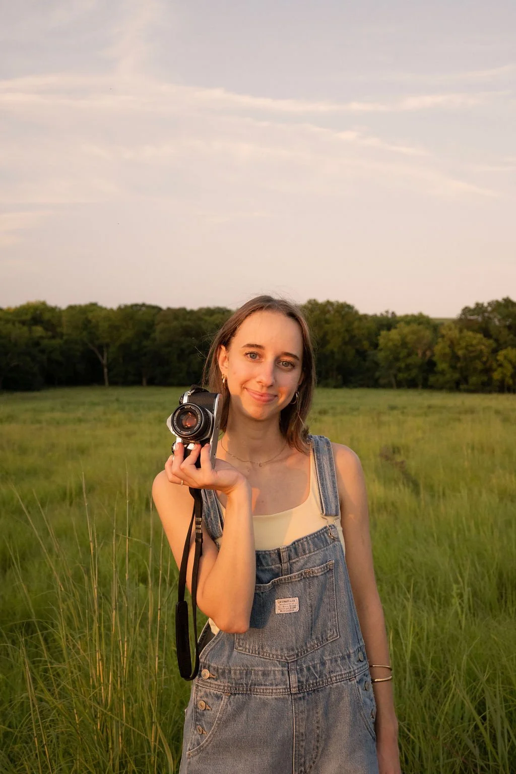 Young woman with shoulder-length brown hair and hoop earrings holding a camera in a grassy field during sunset, with trees in the background and a partly cloudy sky.