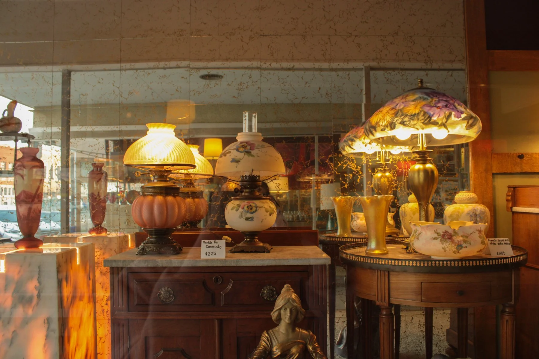 Display of vintage lamps and ceramics inside a store, with a marble-topped table and a carved wood darling mannequin statue in the foreground.