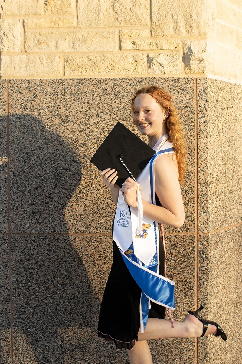 A young woman in a graduation cap and gown holding a diploma and posing with a smile next to a stone wall.