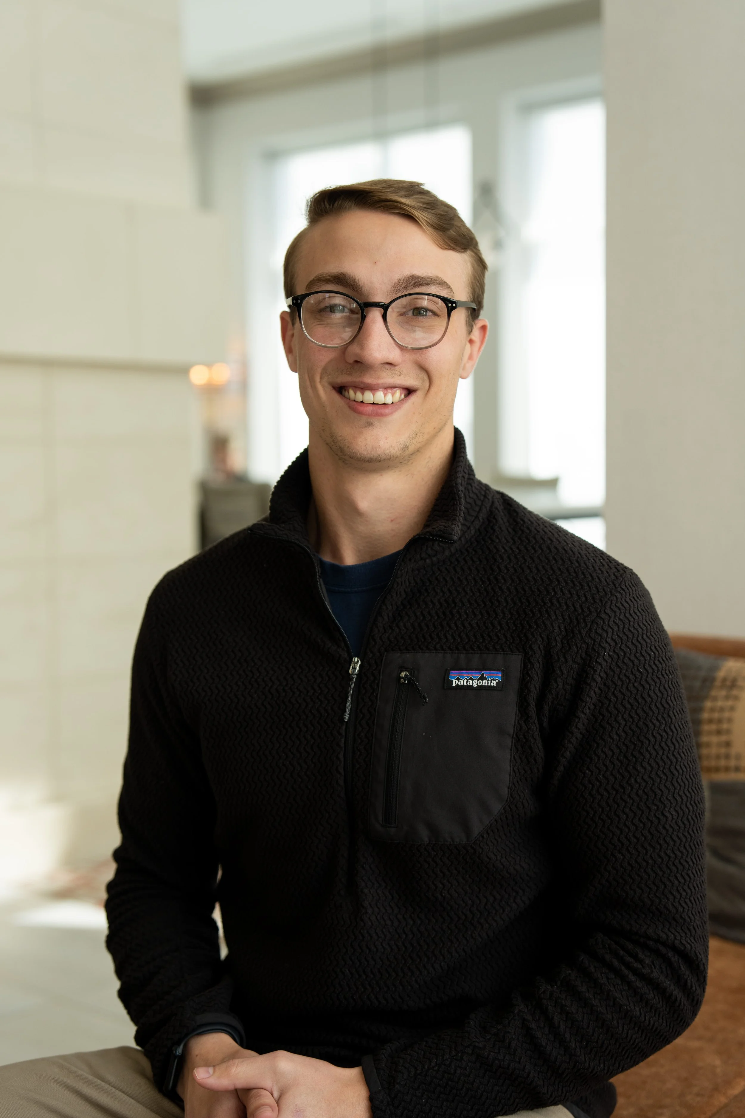 A young man with glasses smiling, wearing a black Patagonia jacket, sitting indoors near a window.