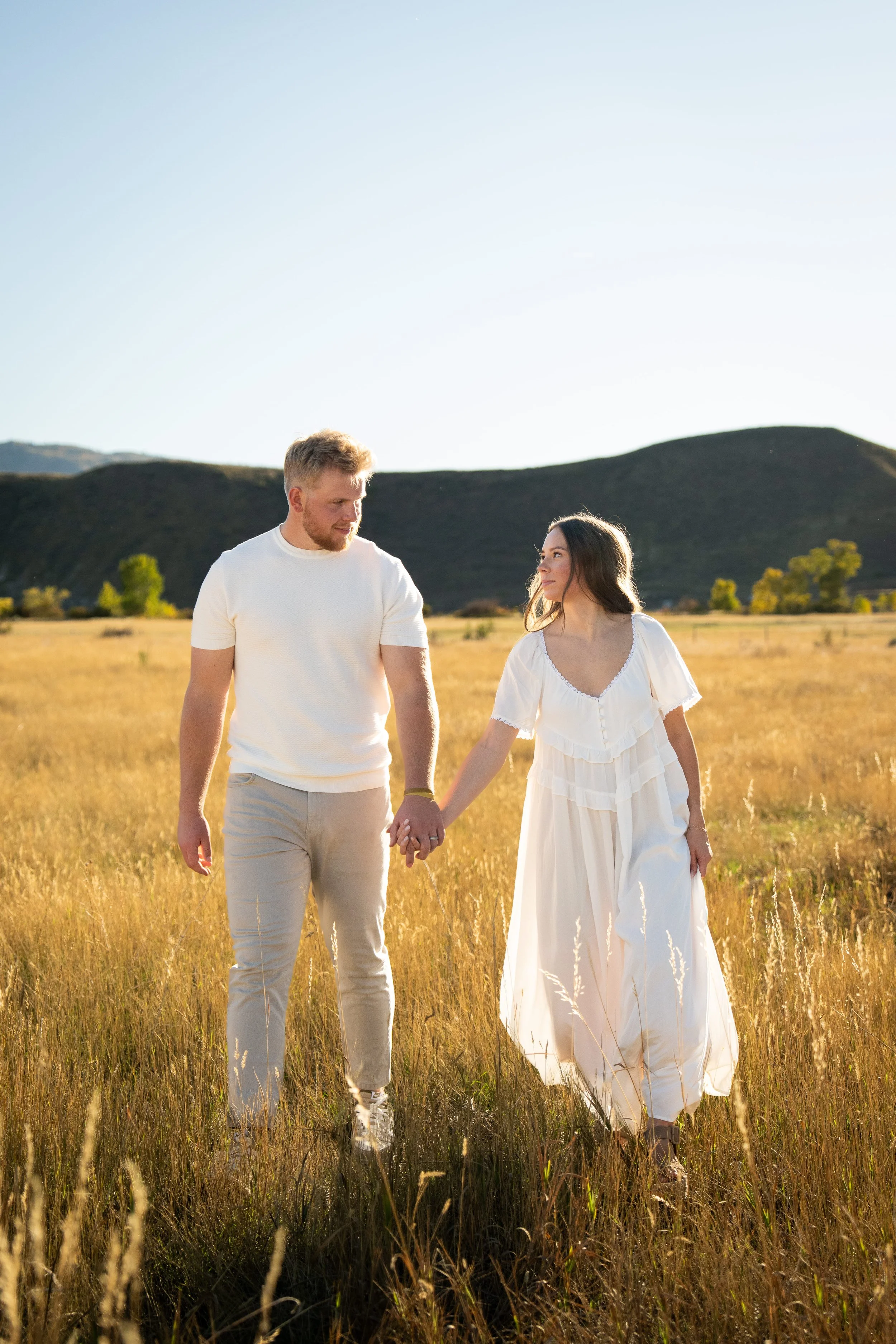 A man and woman holding hands walking through a grassy field with hills in the background at sunset.