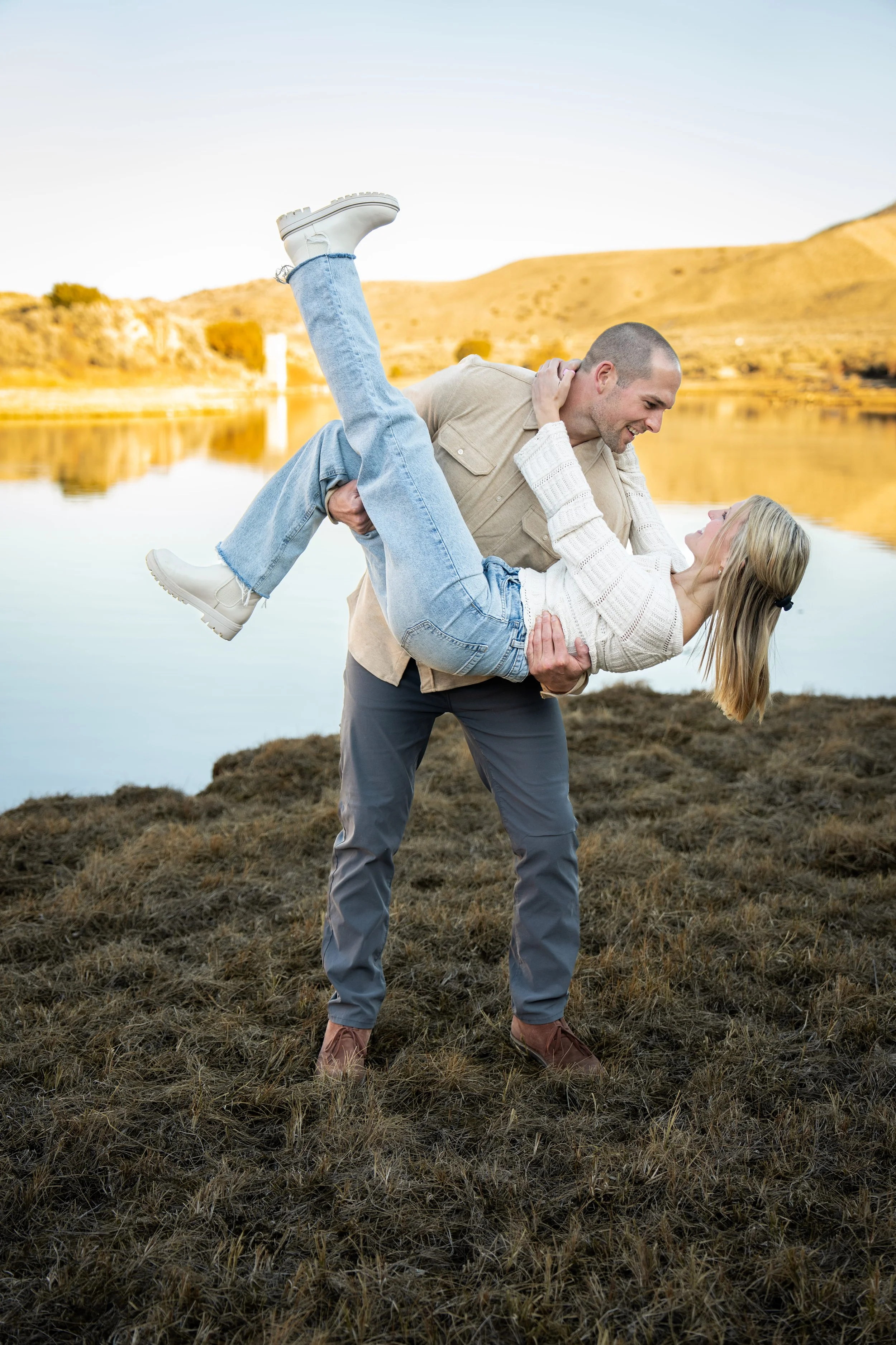 A man and woman outdoors near a lake, with the man lifting the woman in his arms during daytime.