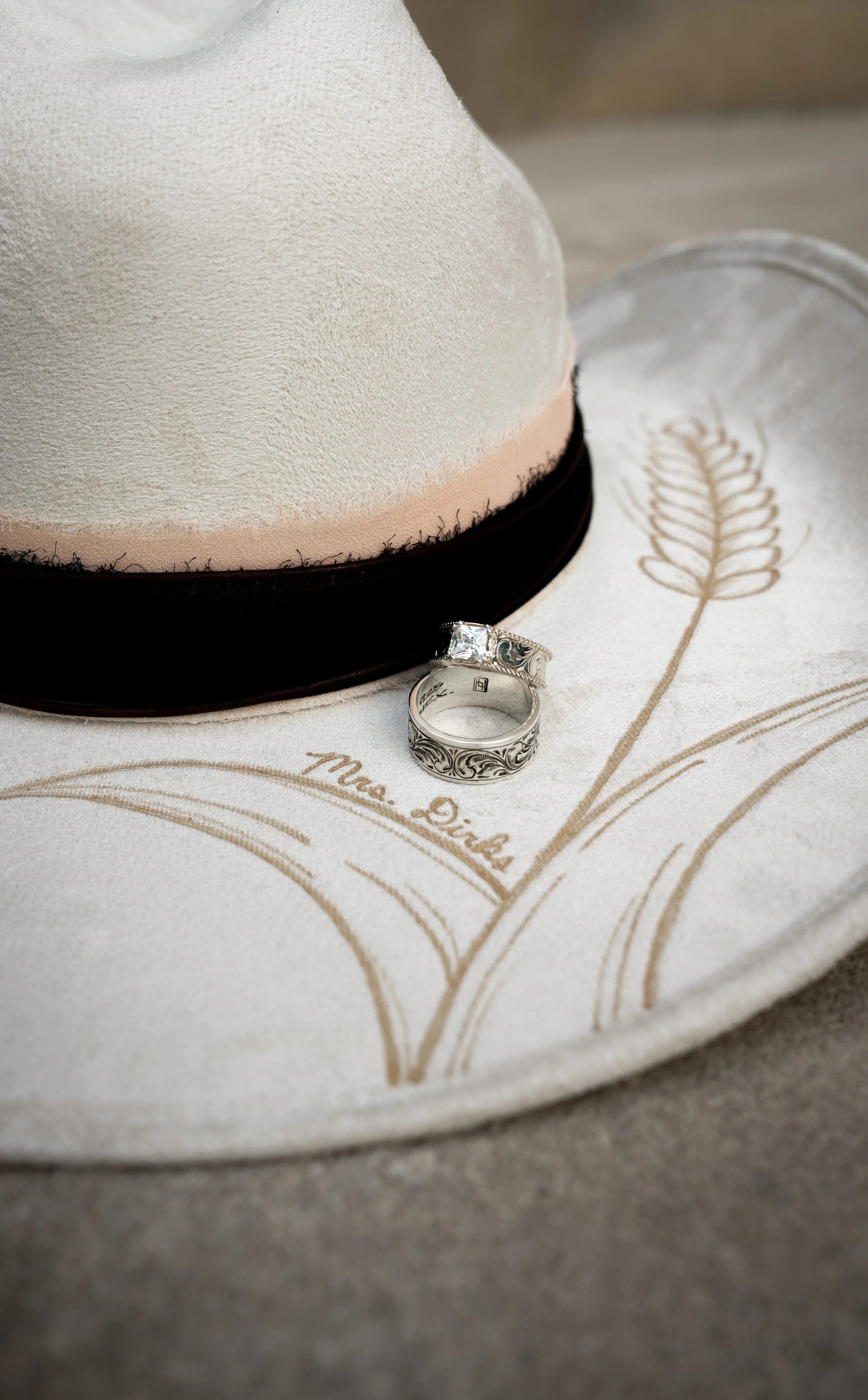 Close-up of two wedding rings placed on a fabric with 'Mrs. Died' inscription, with a hat in the background.