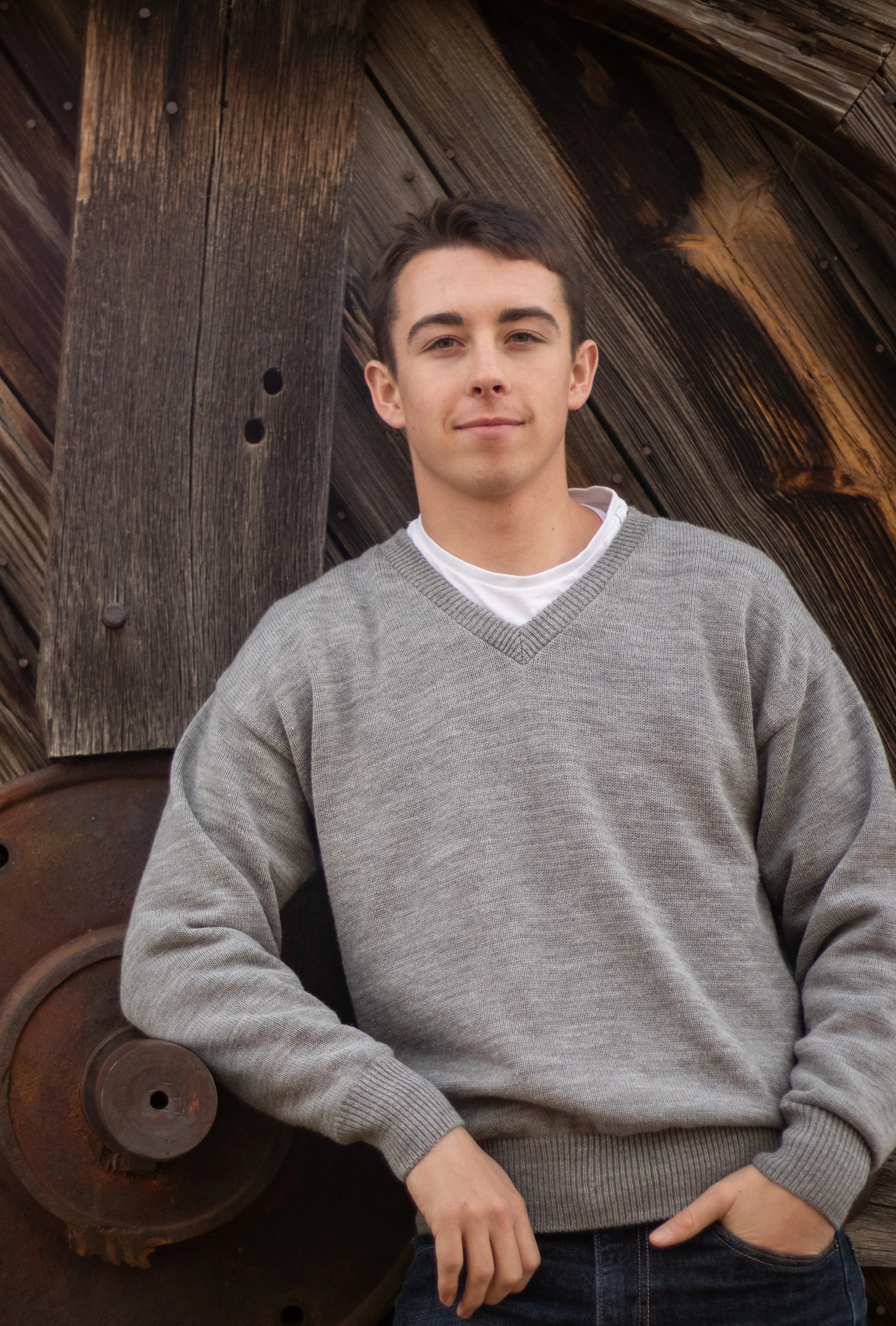 A young man with short dark hair and light skin, wearing a gray V-neck sweater over a white shirt, standing outdoors in front of a weathered wooden structure with rusted metal components. He has a relaxed pose with his left hand in his pocket and a slight smile.