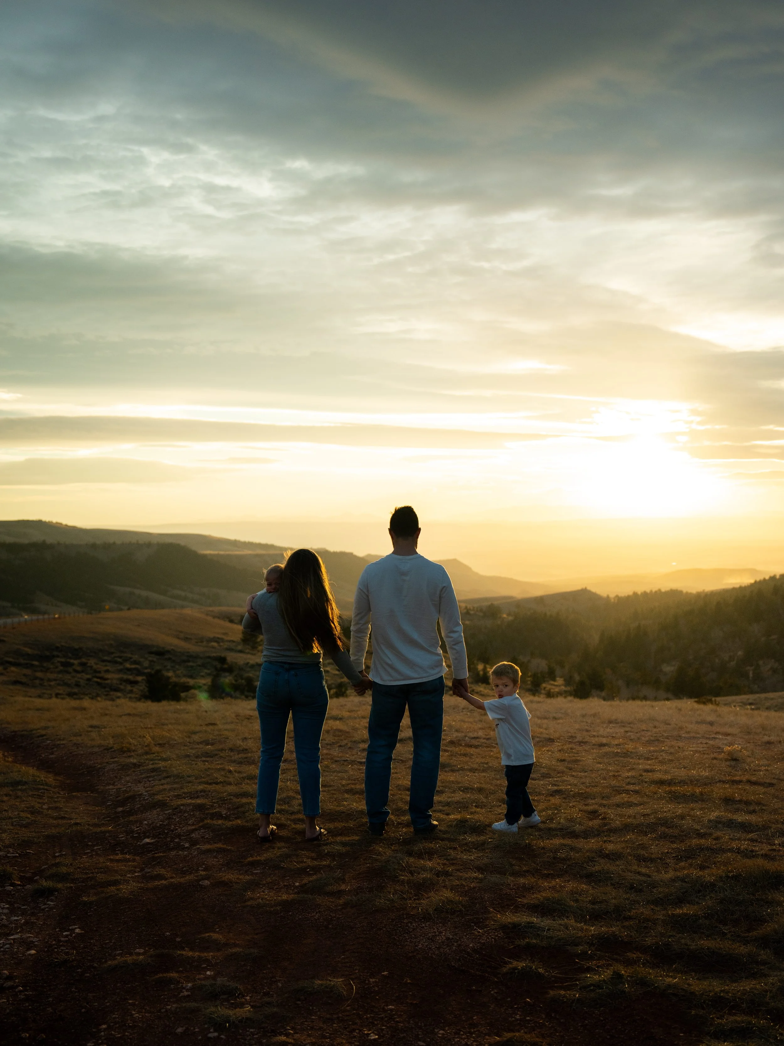 A family of four holding hands and walking in an open field at sunset, with hills and a cloudy sky in the background.