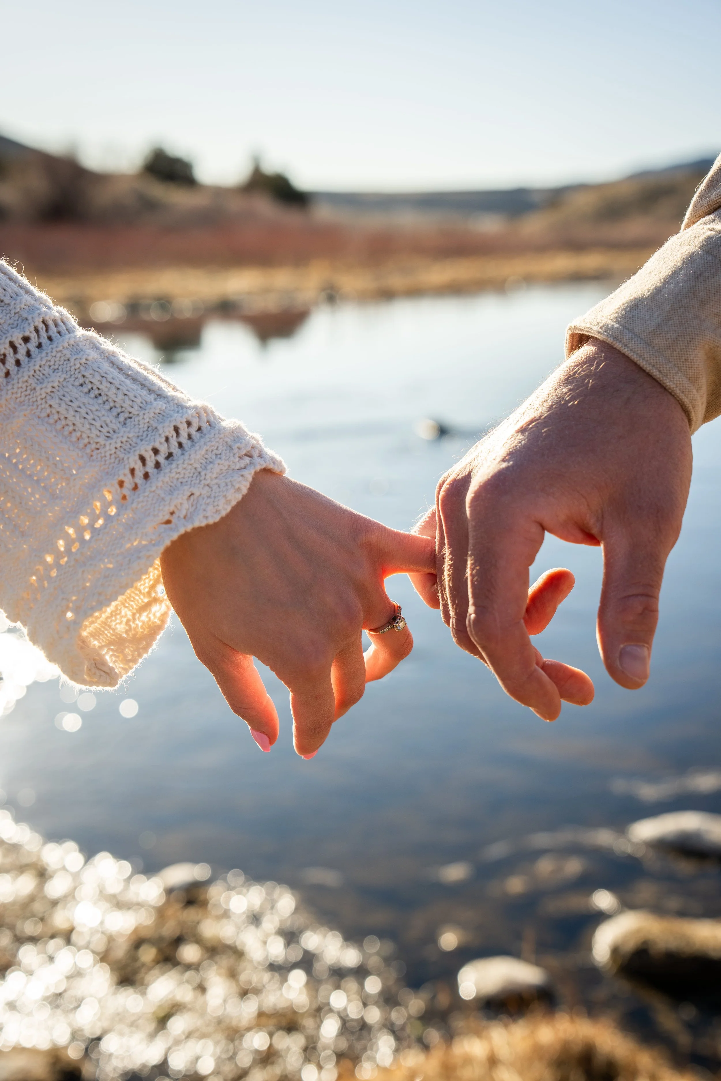 A close-up of two people holding hands near a river, with a blurred natural landscape in the background.