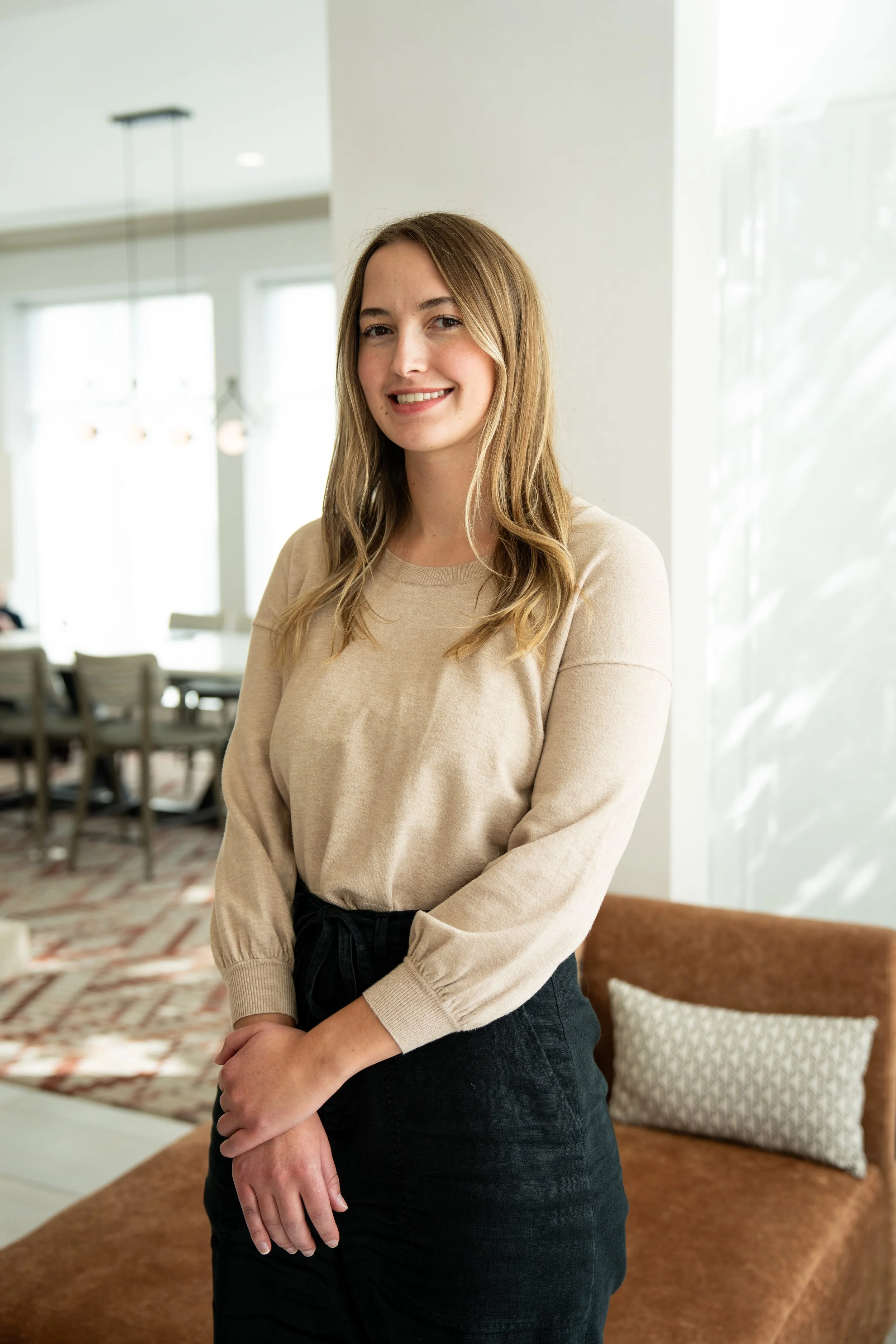 A young woman with long blonde hair, wearing a beige long-sleeve top and black skirt, standing in a bright, modern indoor space with large windows and a brown couch in the background.