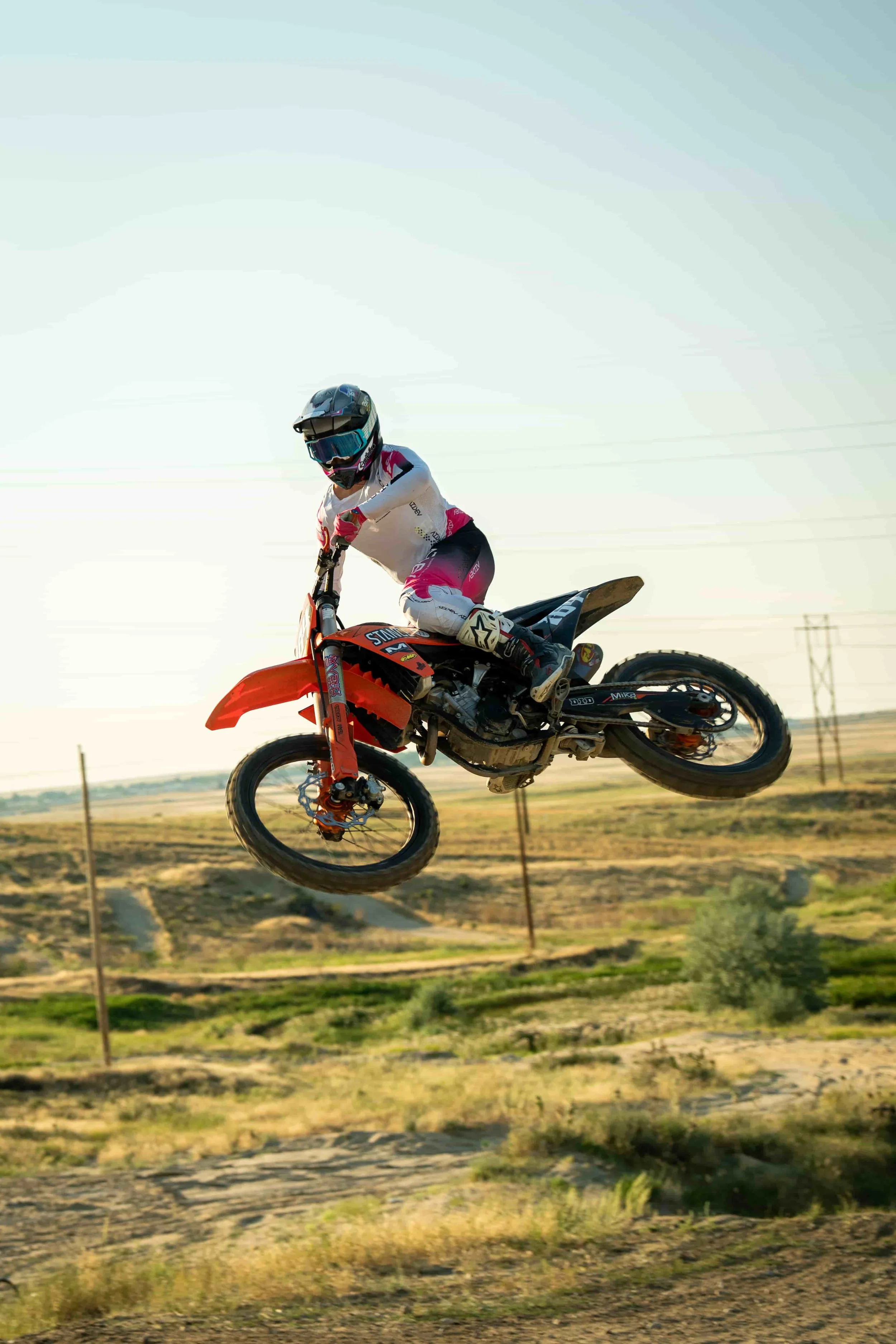 A motocross rider wearing a black helmet, white and pink gear, mid-air on an orange dirt bike during a jump in an outdoor dirt track setting.