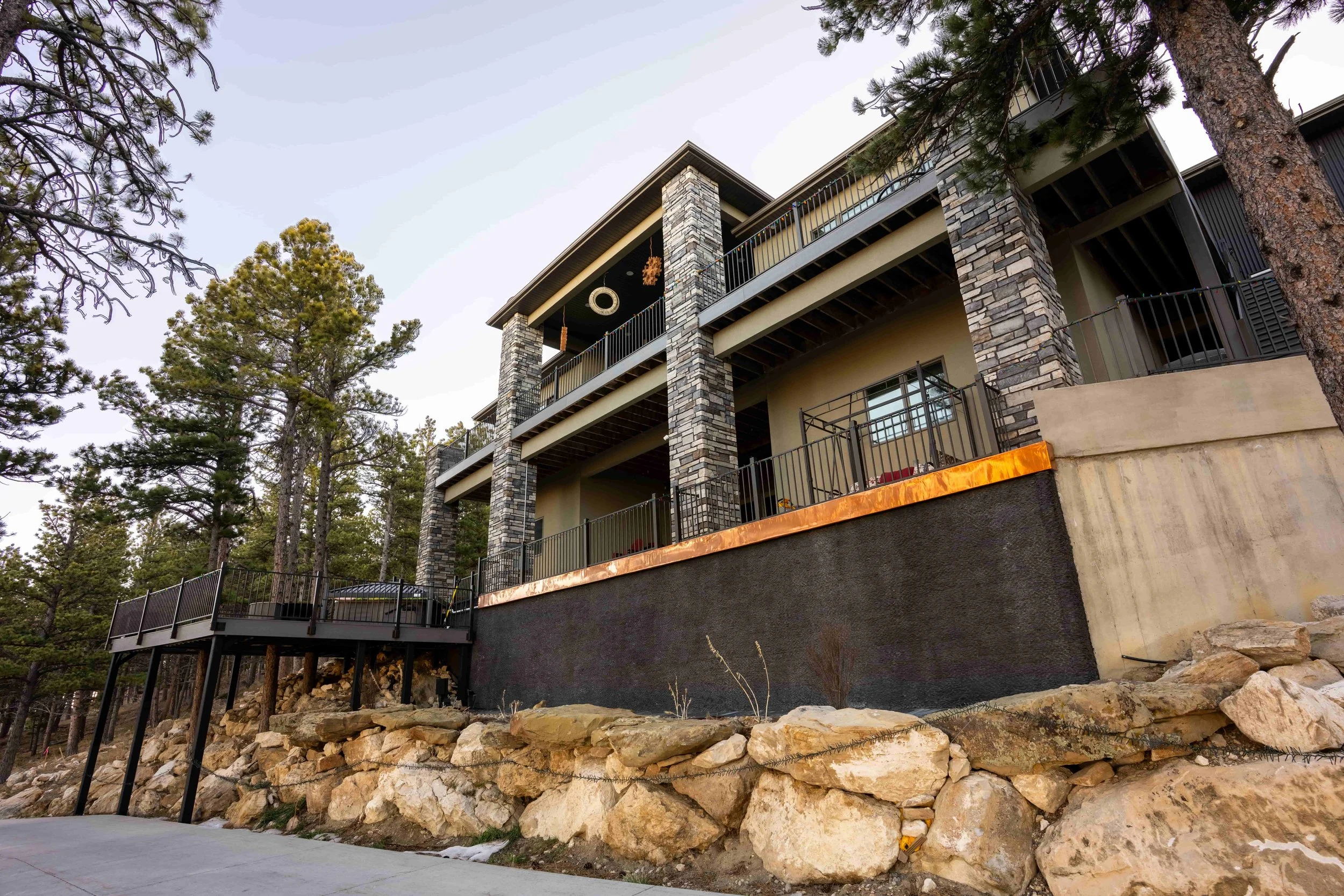 Multi-story modern house built on a rocky hillside with stone columns, metal railings, and outdoor balconies, surrounded by tall pine trees.