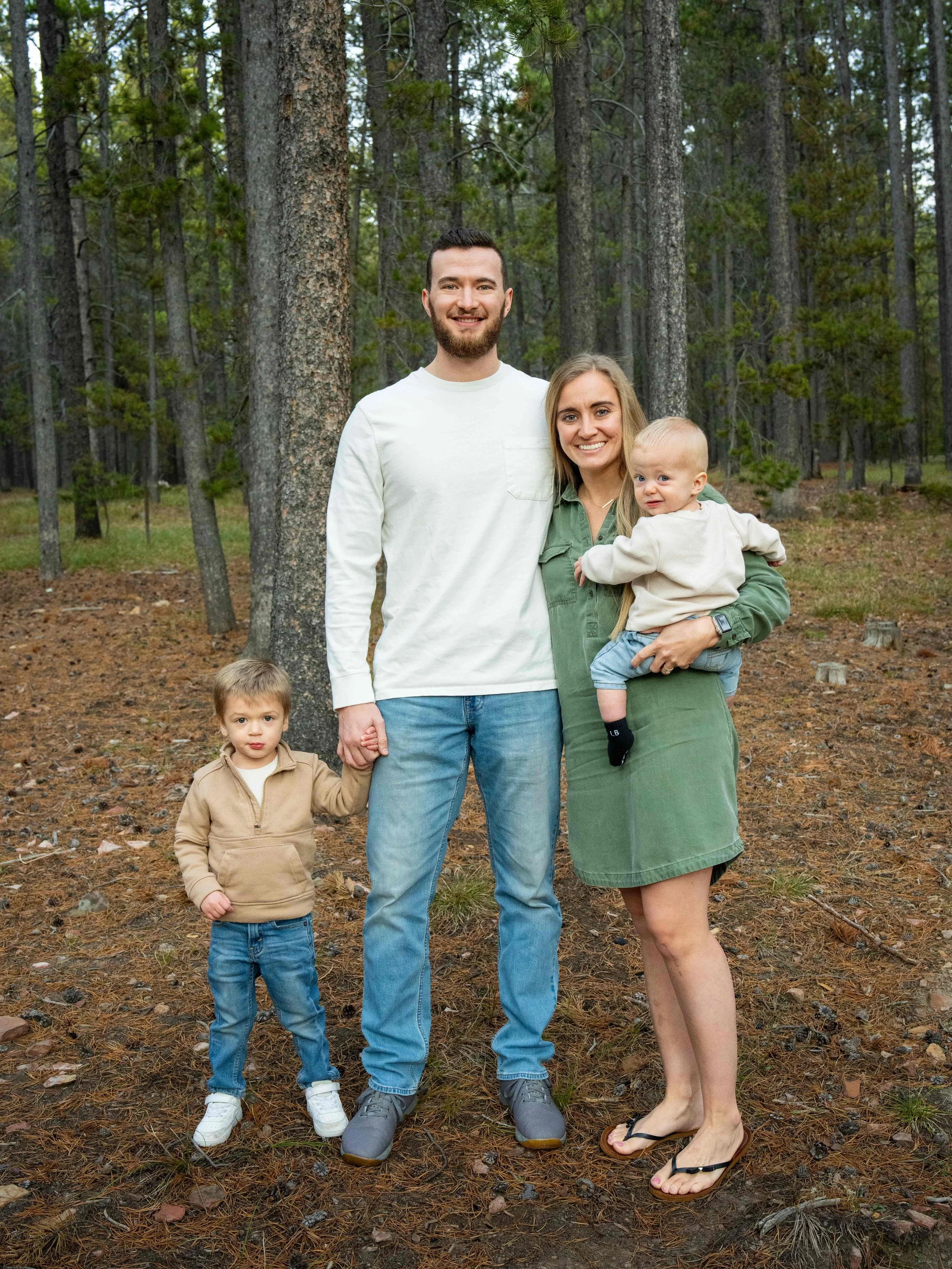 A family of four standing outdoors in a wooded area, holding hands and smiling.