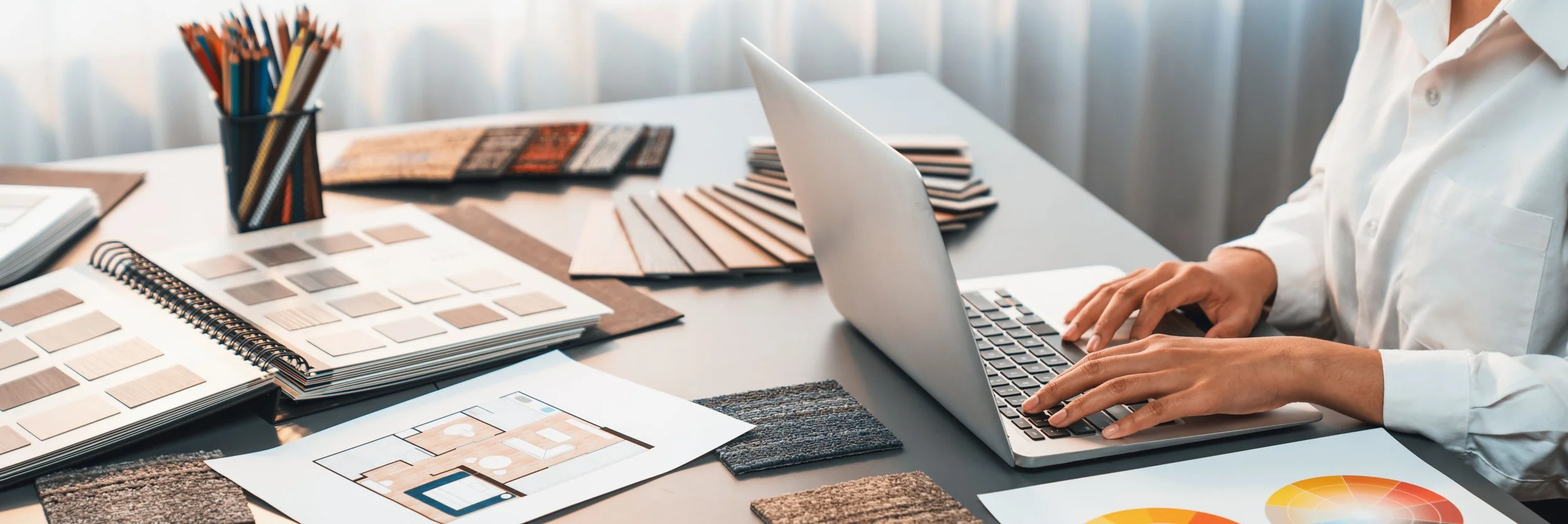 Person working on a laptop surrounded by interior design samples, fabric swatches, color palettes, and floor plans, in a well-lit room.