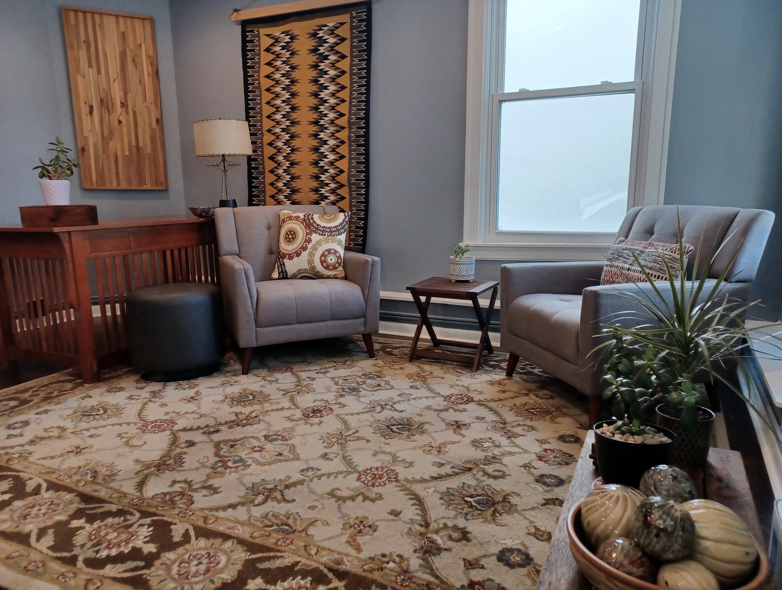 Living room with two armchairs, side table with plant, decorated woven wall hanging, wooden cabinet, modern lamp, patterned rug, and potted plants near window.