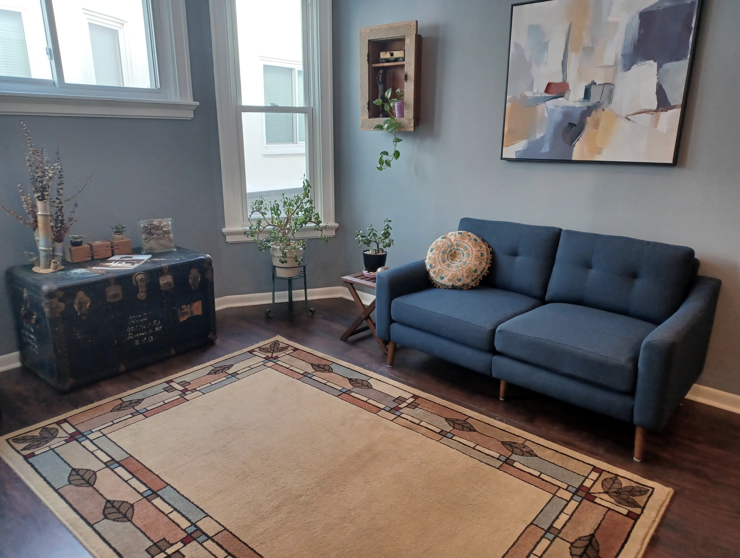 A cozy living room corner with a blue sofa, colorful round pillow, wooden side table with plants, vintage trunk with decorative items, a large abstract painting on the wall, and a beige patterned rug on dark hardwood floor, illuminated by natural lig