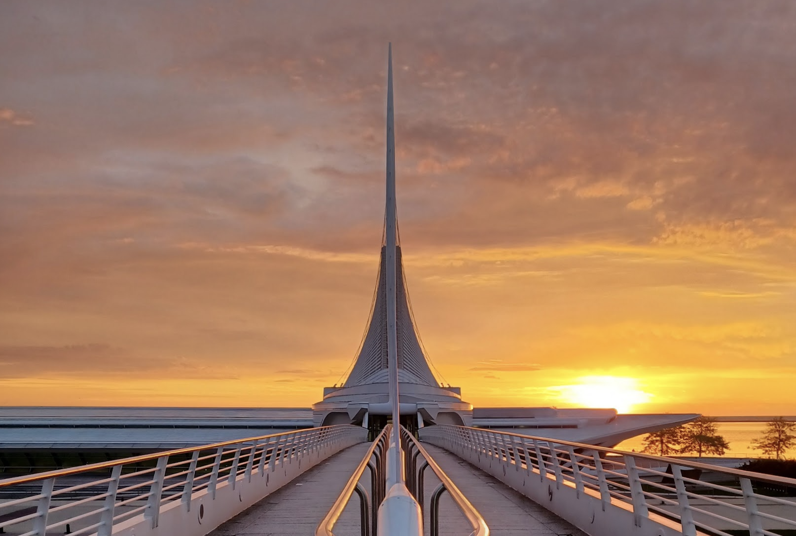 A modern architectural bridge or walkway with a futuristic design, leading towards a central tower with a sharp spire, during sunset with a colorful sky.