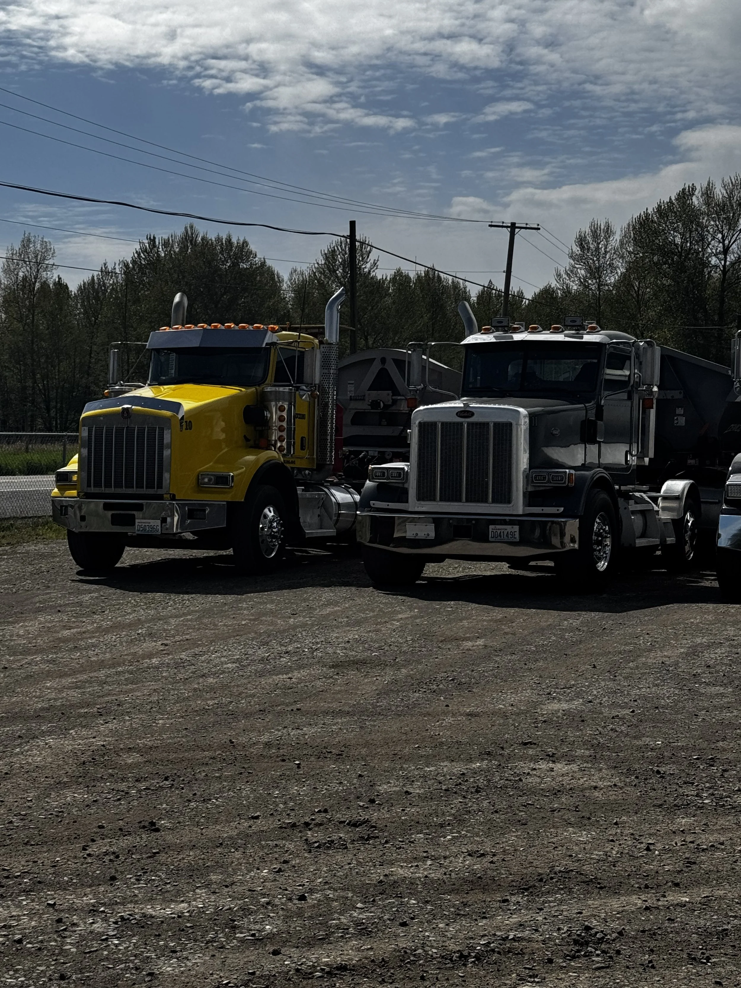 Two semi-trucks parked on a gravel lot under a partly cloudy sky, one yellow and one silver.