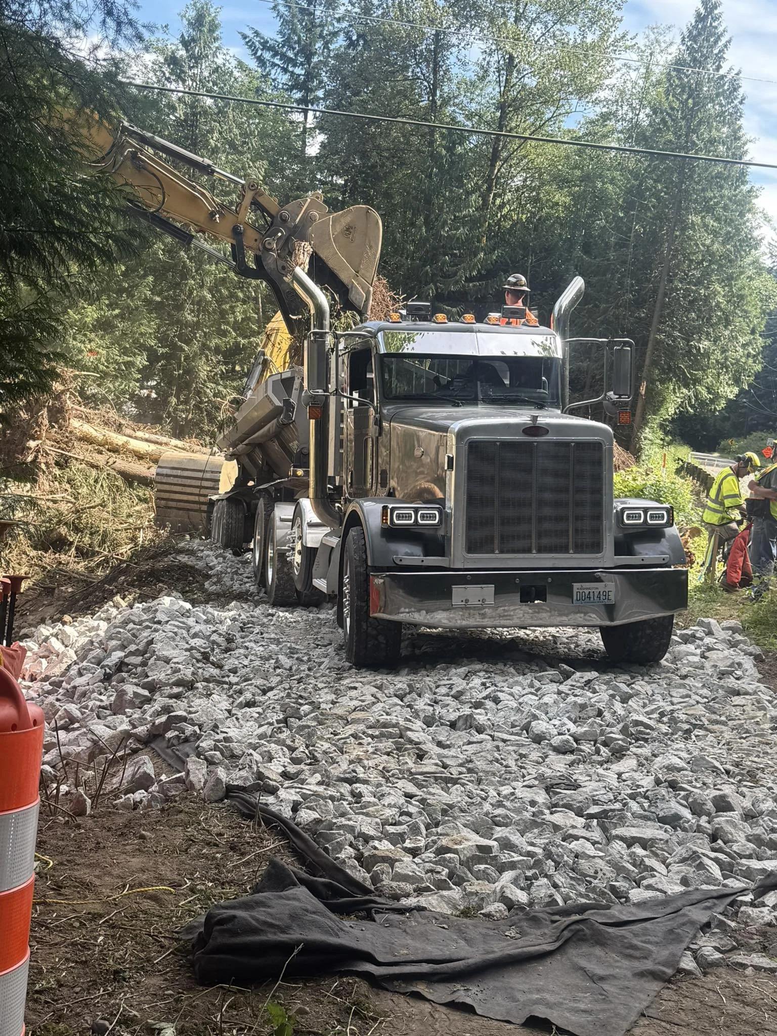 A large construction truck with a yellow excavator arm working on a rocky unfinished road, surrounded by green trees and construction workers in safety vests.