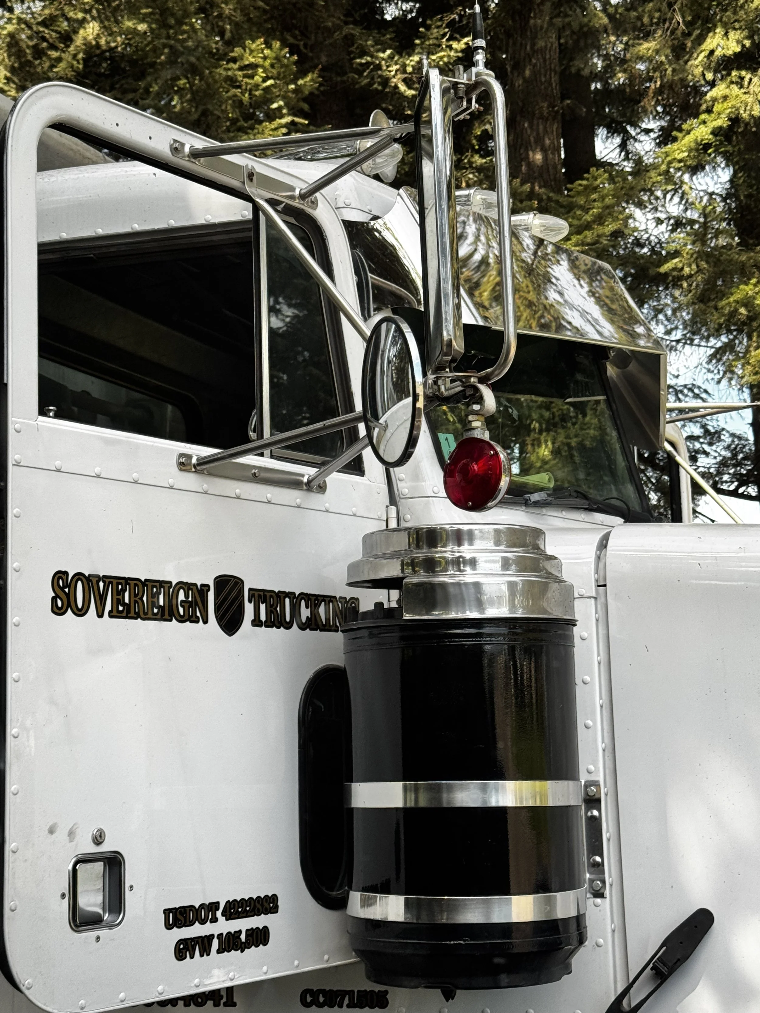 Close-up of a large white semi-truck with chrome details, black fuel tank, and a side mirror, featuring the text 'SOVEREIGN TRUCKING' on the door, set against a background of trees.