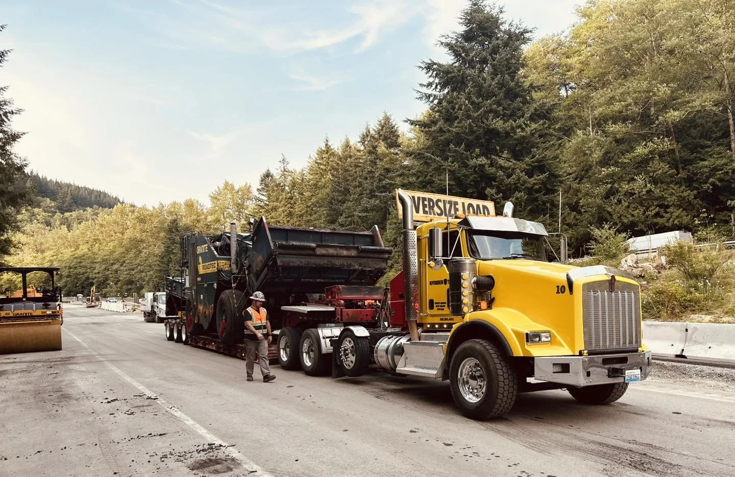 A large yellow construction truck on a road, with a worker in an orange vest and hard hat standing nearby, surrounded by trees and construction equipment.