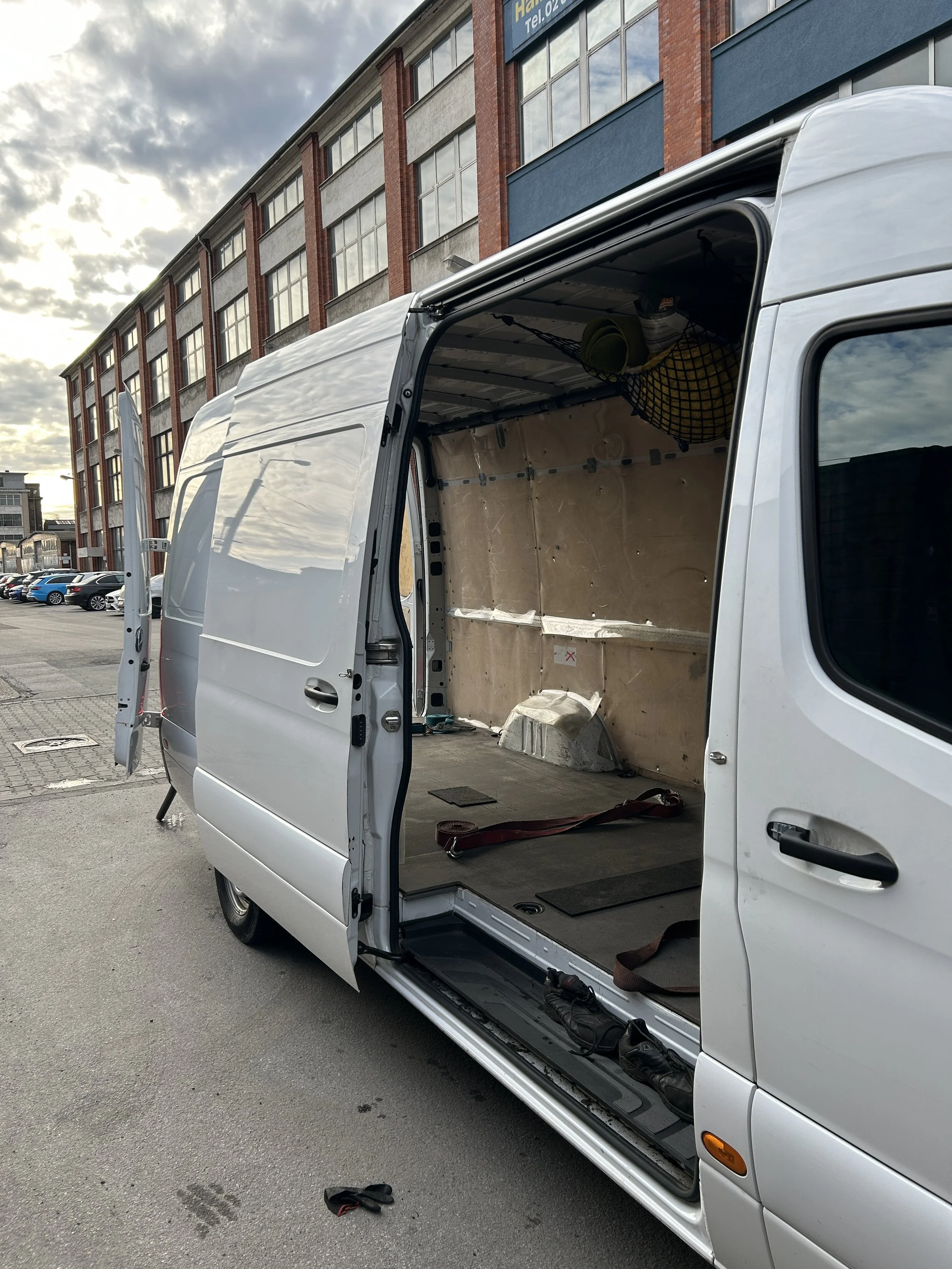 Leere weiße Lieferwagen mit offener Seitentür, im Hintergrund ein mehrstöckiges Gebäude mit großen Fenstern und Parkplatz.