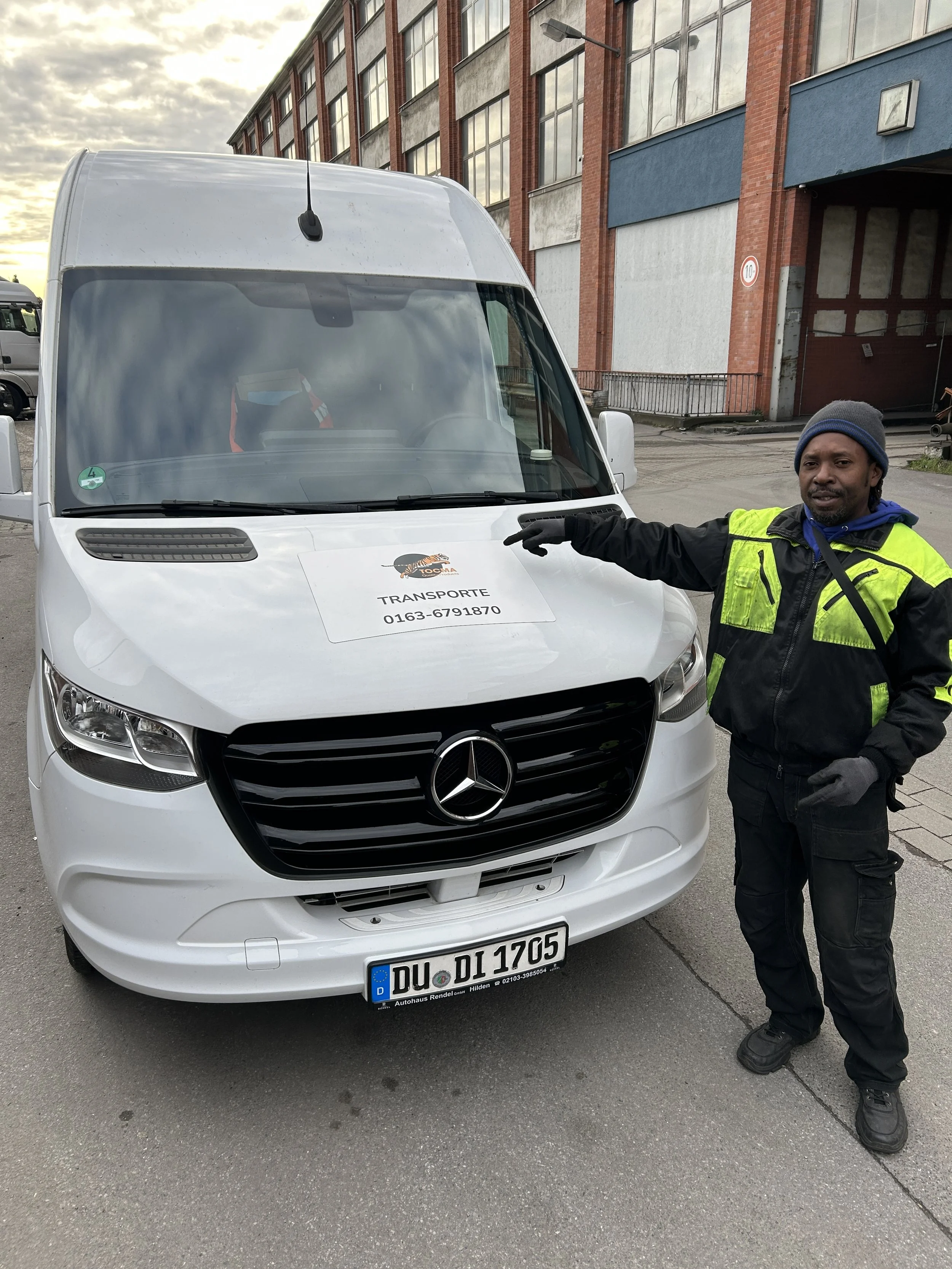 Ein Mann in einer schwarzen Jacke mit gelben Akzenten steht neben einem weißen Mercedes-Transporter auf einer Straße. Er zeigt auf das Fahrzeug, das eine rote Warnweste im Innenraum hat. Auf der Windschutzscheibe ist ein Schild mit Fahrzeugdetails und Telefonnummer. Das Fahrzeug hat ein deutsches Nummernschild.