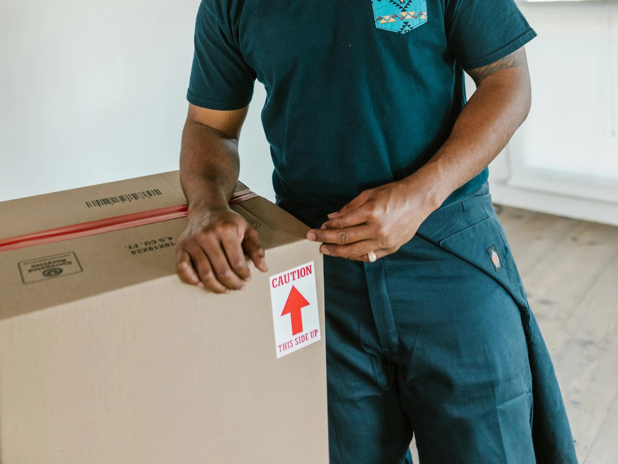 Eine Person beim Versand eines Pakets, hält eine große Kartonschachtel, die ein Warnetikett mit einem roten Pfeil und der Aufschrift "Vorsicht, diese Seite nach oben" trägt.