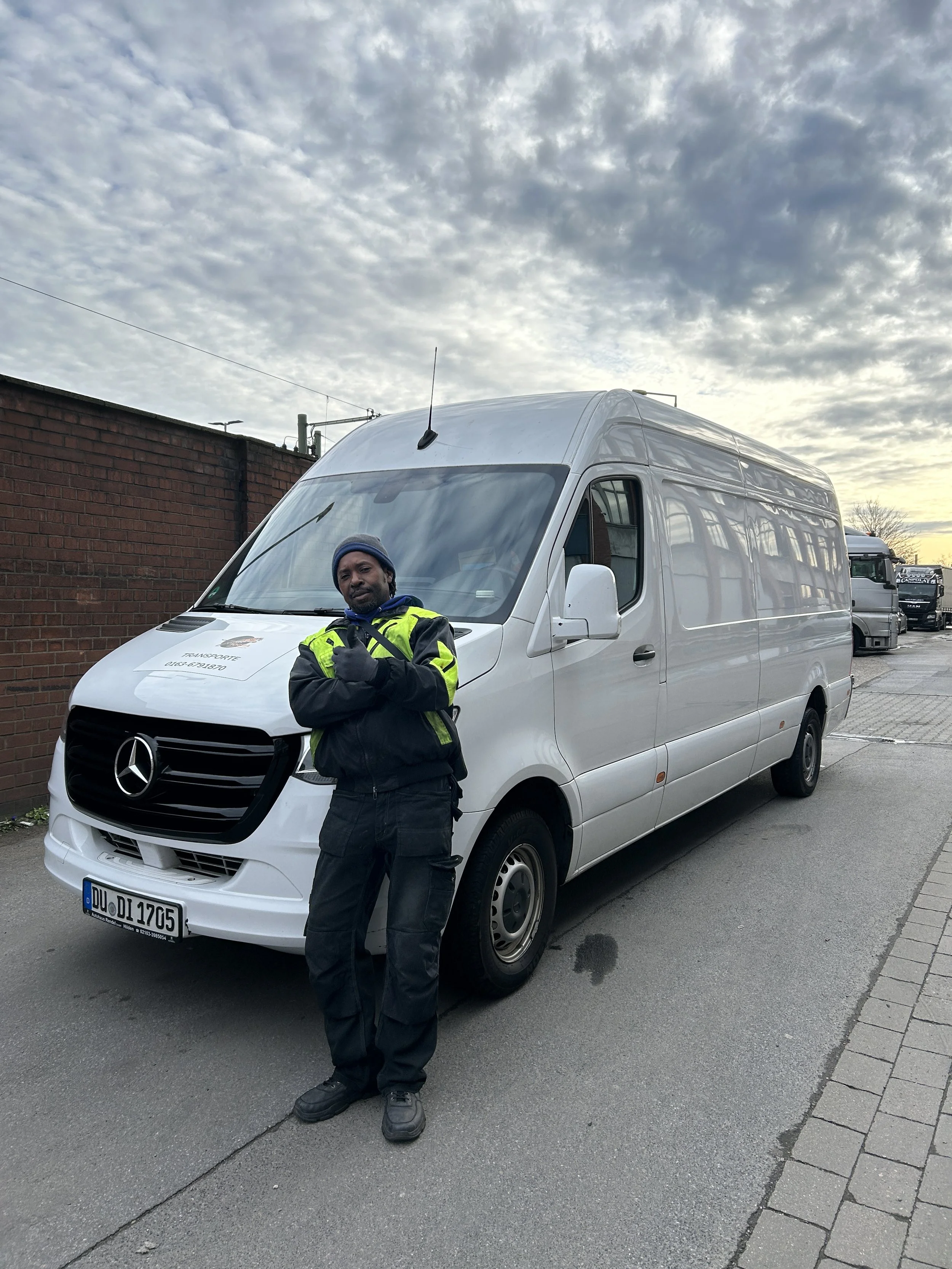 Ein Mann steht vor einem weißen Mercedes-Benz Transporter auf einem Stadtraum. Im Hintergrund ist ein Brickwall und mehrere Lkw. Der Mann trägt eine schwarze Jacke mit Neon-Grün-Haltes und macht ein Daumen-hoch-Geste.