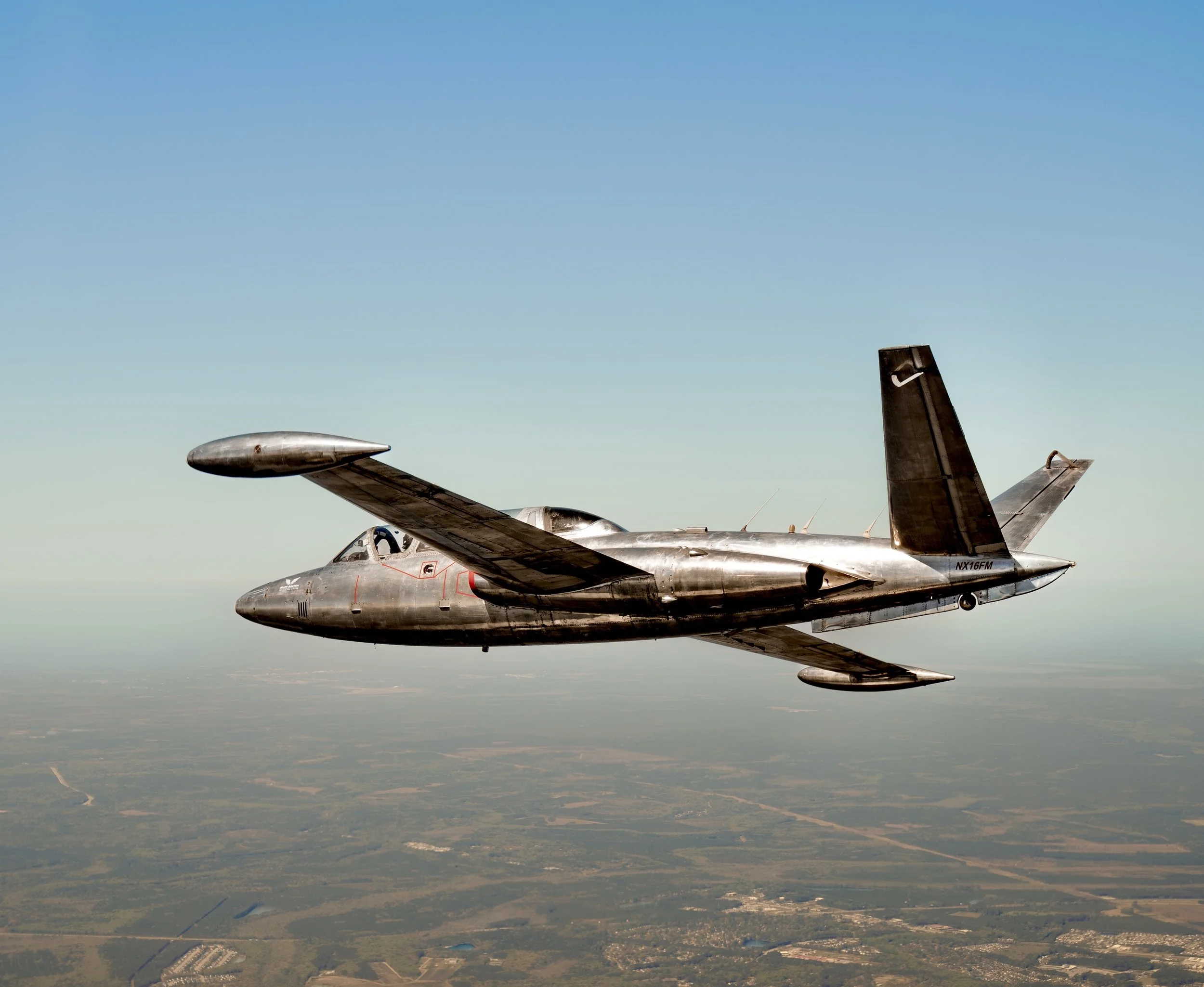A futuristic silver spaceship flying high above the ground with fields and roads below and a clear blue sky.