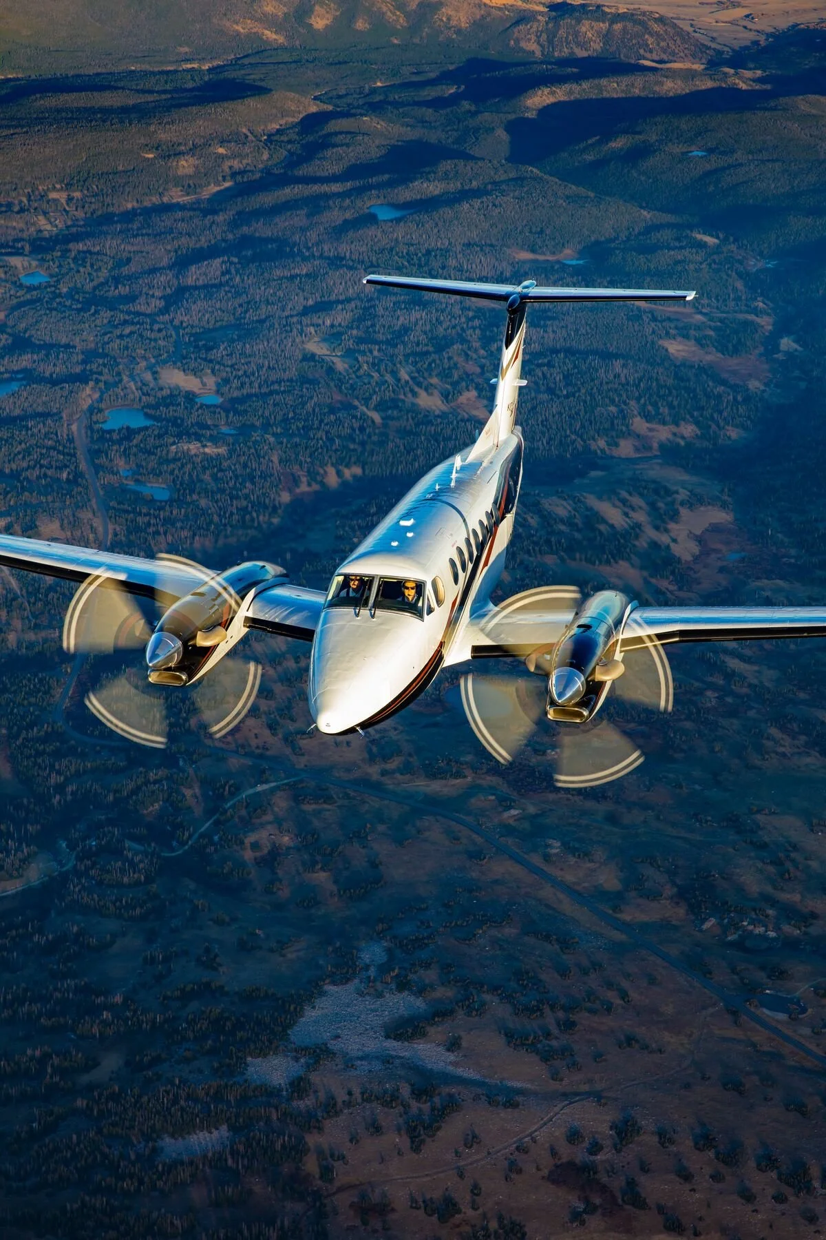 A twin-engine propeller airplane flying over a landscape with forests and lakes, at sunset.