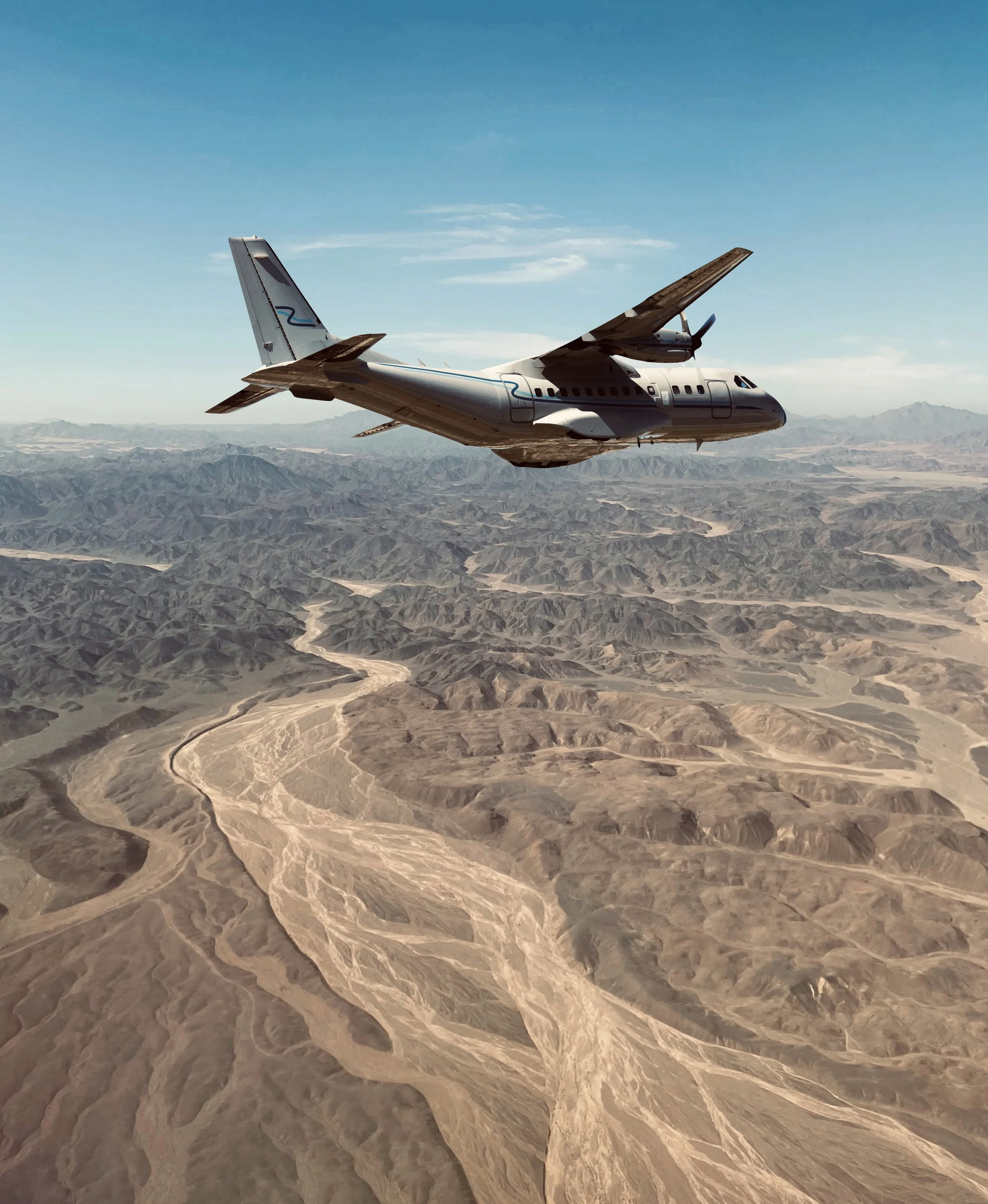 A silver airplane flying above a desert landscape with mountains and winding rivers under a clear sky.