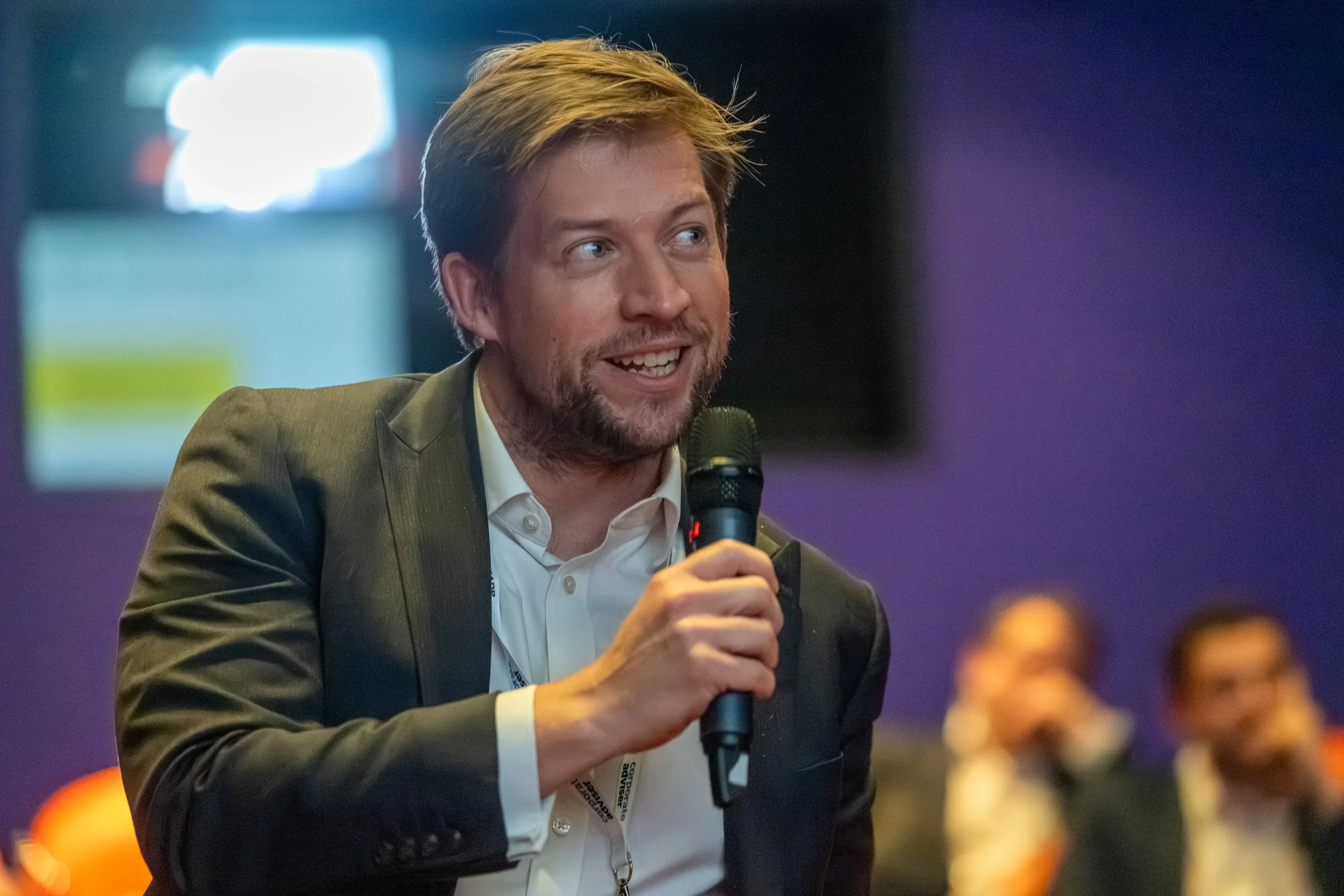 A man in a suit speaking into a microphone during a conference or event, with people seated in the background.