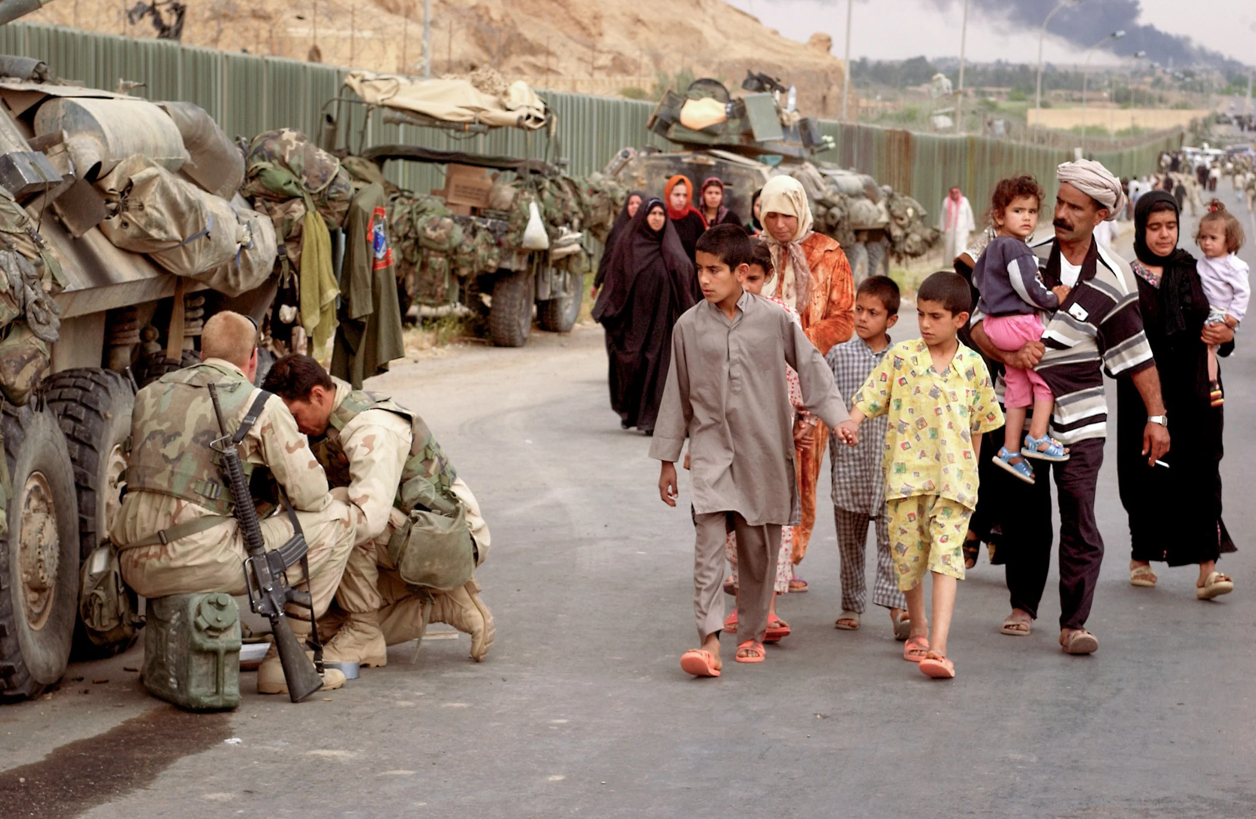 Military personnel by their vehicles, as a family of civilians, including children and women, walk by on a rural road with more military vehicles and smoke rising in the background.