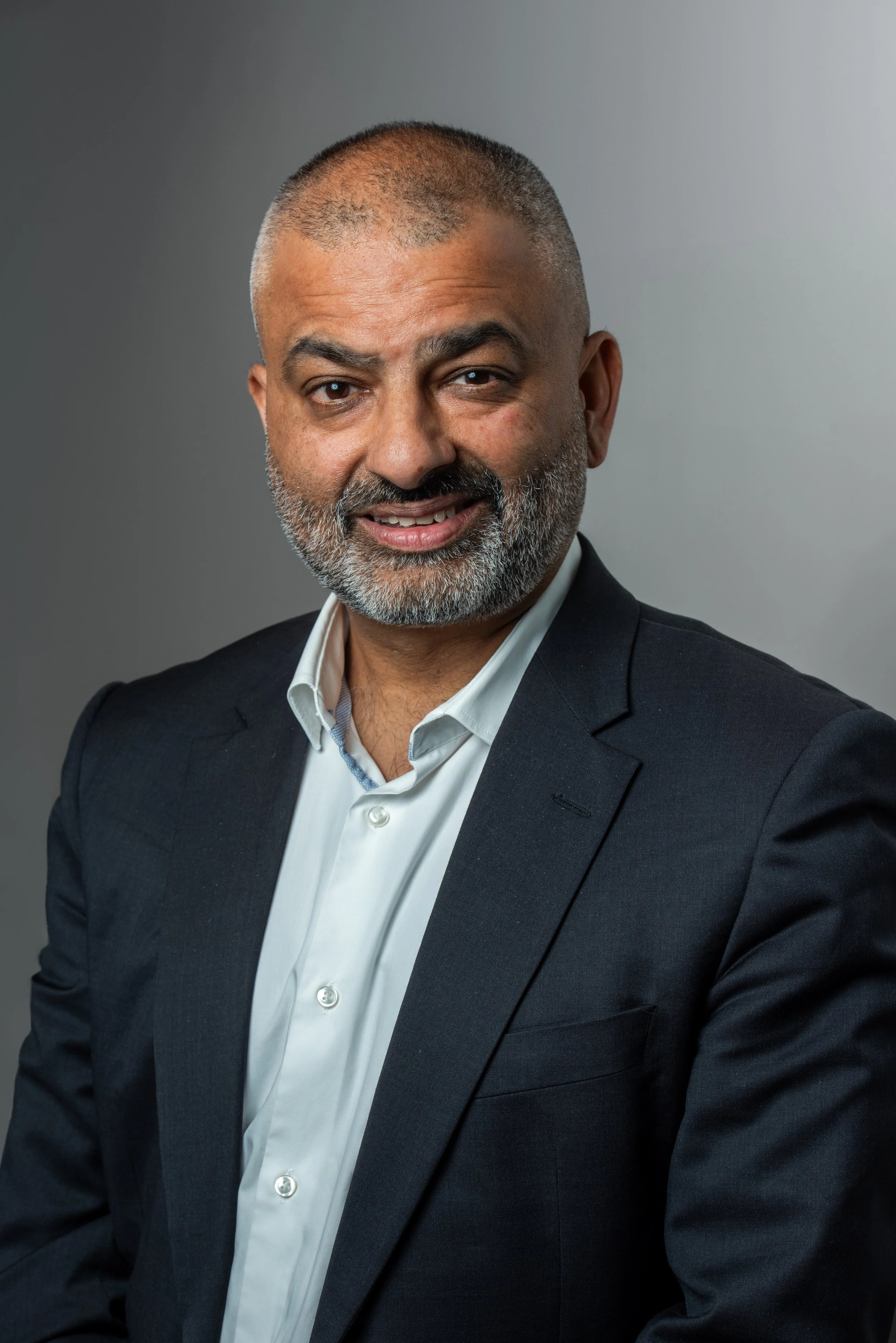 A middle-aged man with a beard and short hair, wearing a dark suit jacket and white dress shirt, smiling at the camera against a plain gray background.
