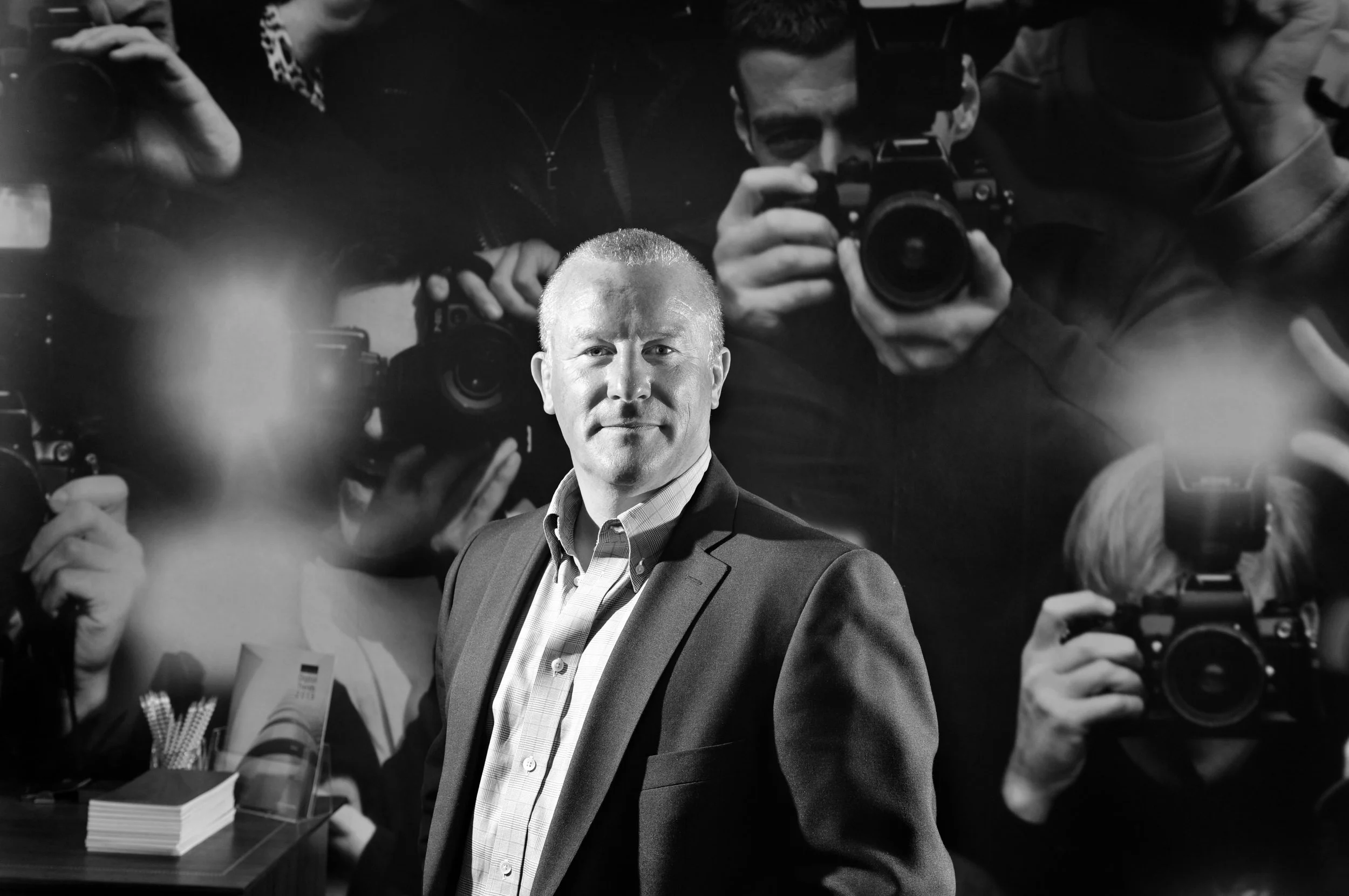 A man in a suit stands in front of a dark background with photographers taking pictures of him, some using cameras with large lenses. The image is in black and white.