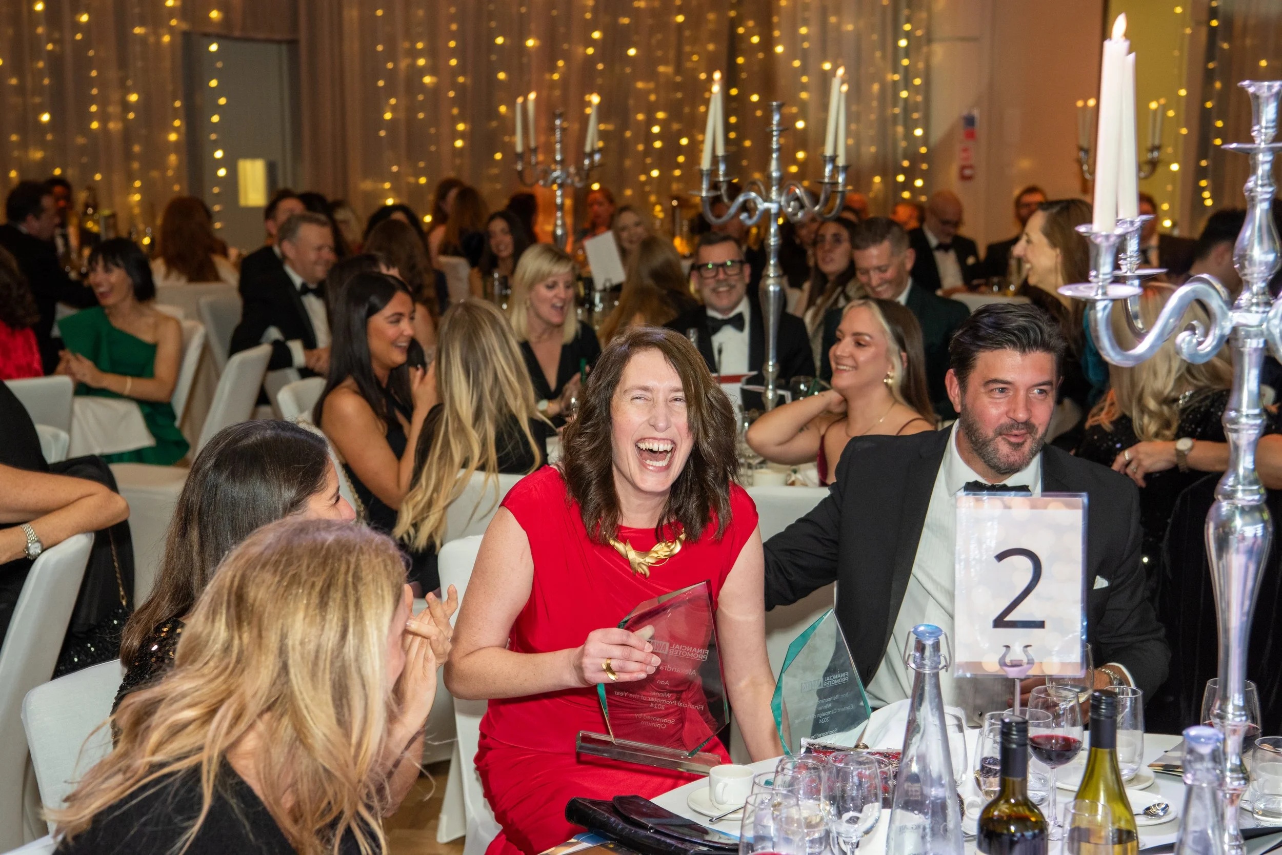 People dressed in formal attire, sitting at tables, attending a festive event or gala, with some smiling and laughing, with string lights and candles in the background.