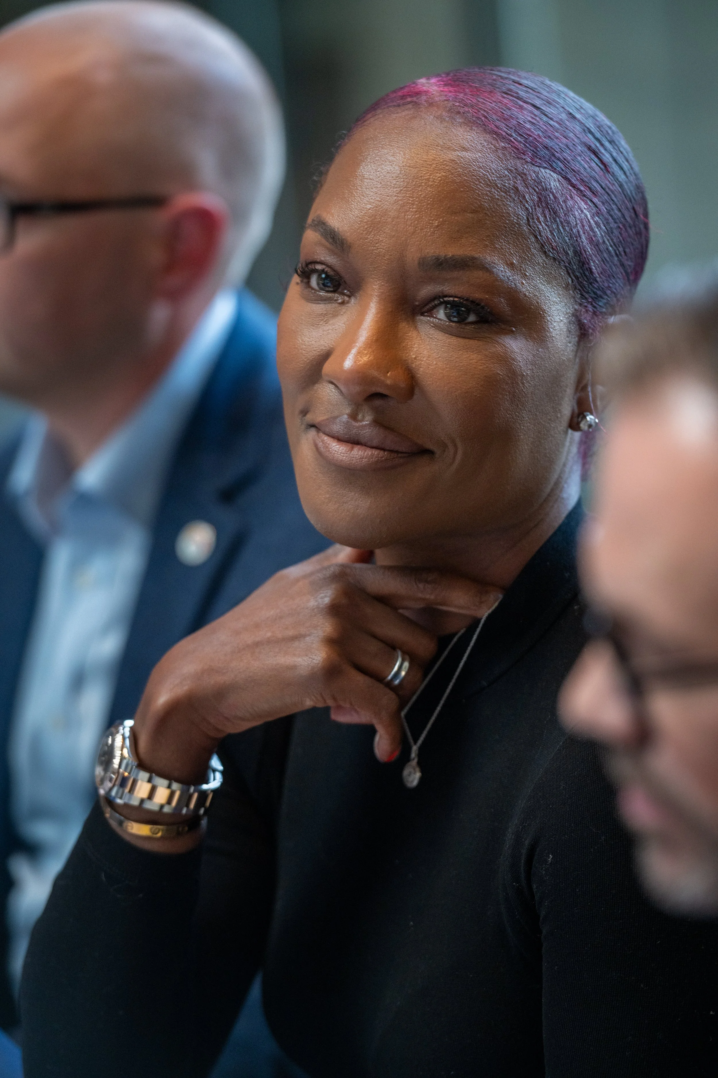 An African American woman in a black top sitting at a meeting or event, with a thoughtful expression, hand on her neck, wearing a watch, necklace, and earrings.