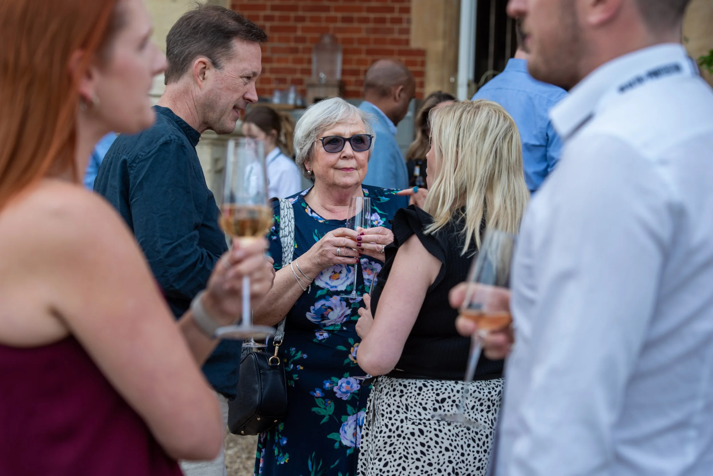 Group of people at an outdoor social gathering, holding glasses of wine, conversing, with a brick building in the background.