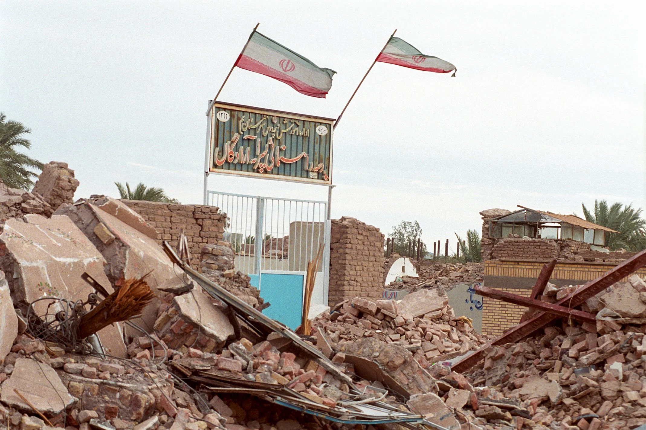 Ruins of a destroyed building with debris and rubble, two Iranian flags on a signboard, and palm trees in the background.