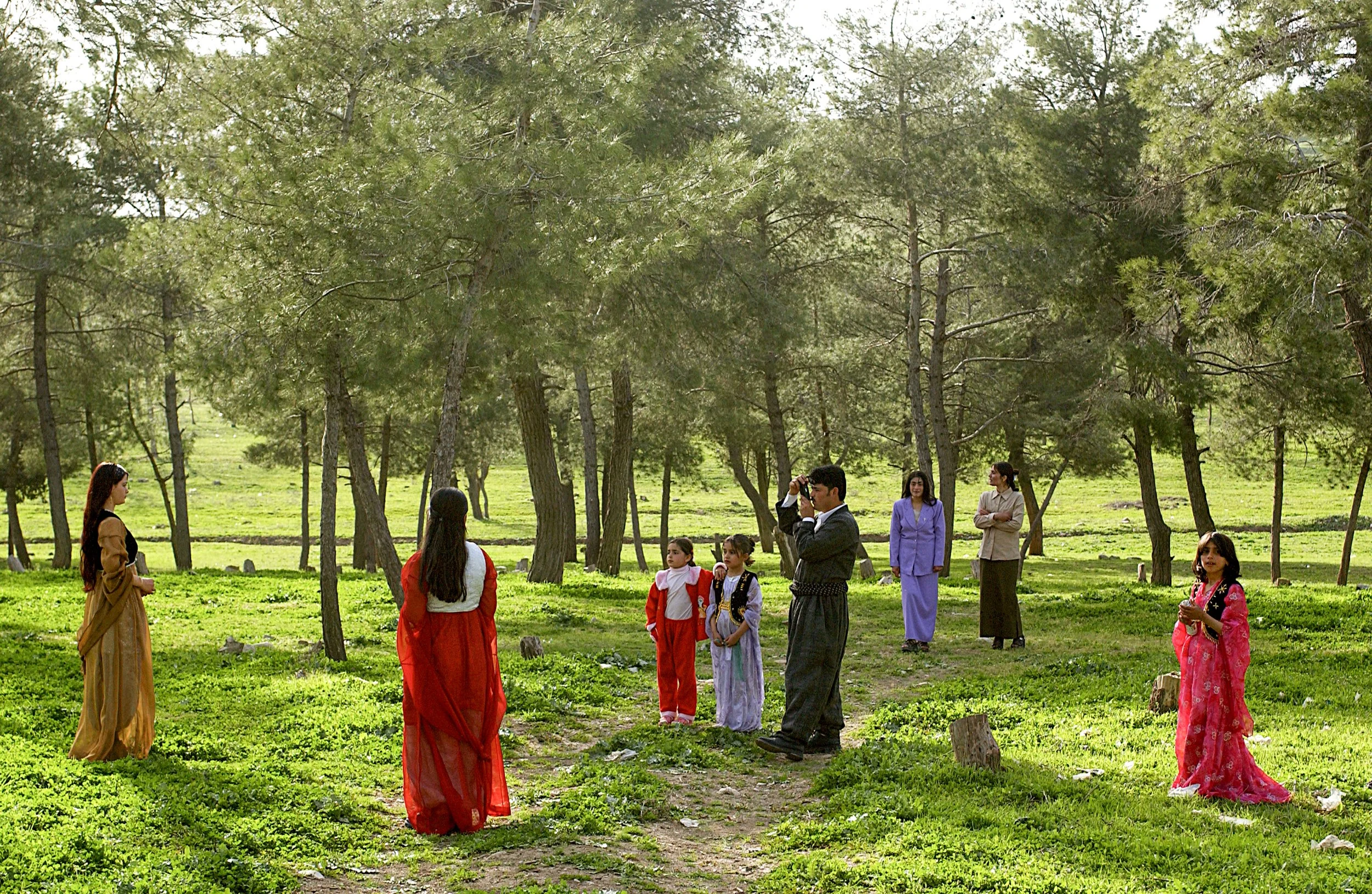 People in traditional clothing gathering in a green park with tall trees.