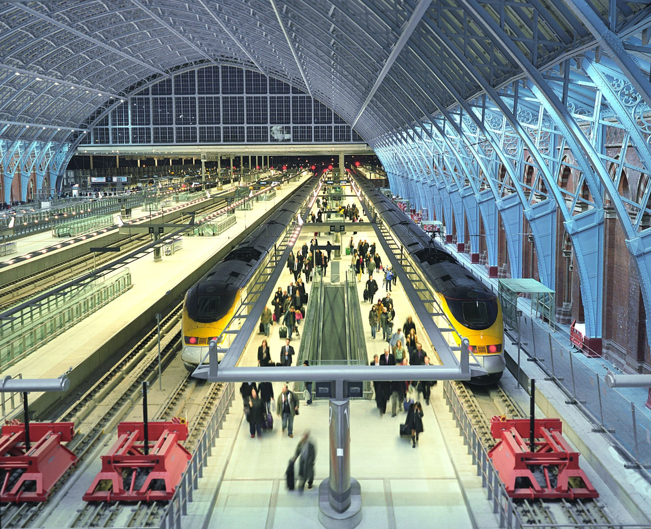 Inside St Pancras International train station with a high arched glass and steel roof, multiple train tracks, and a platform with passengers waiting and walking, two yellow and black trains on either side.