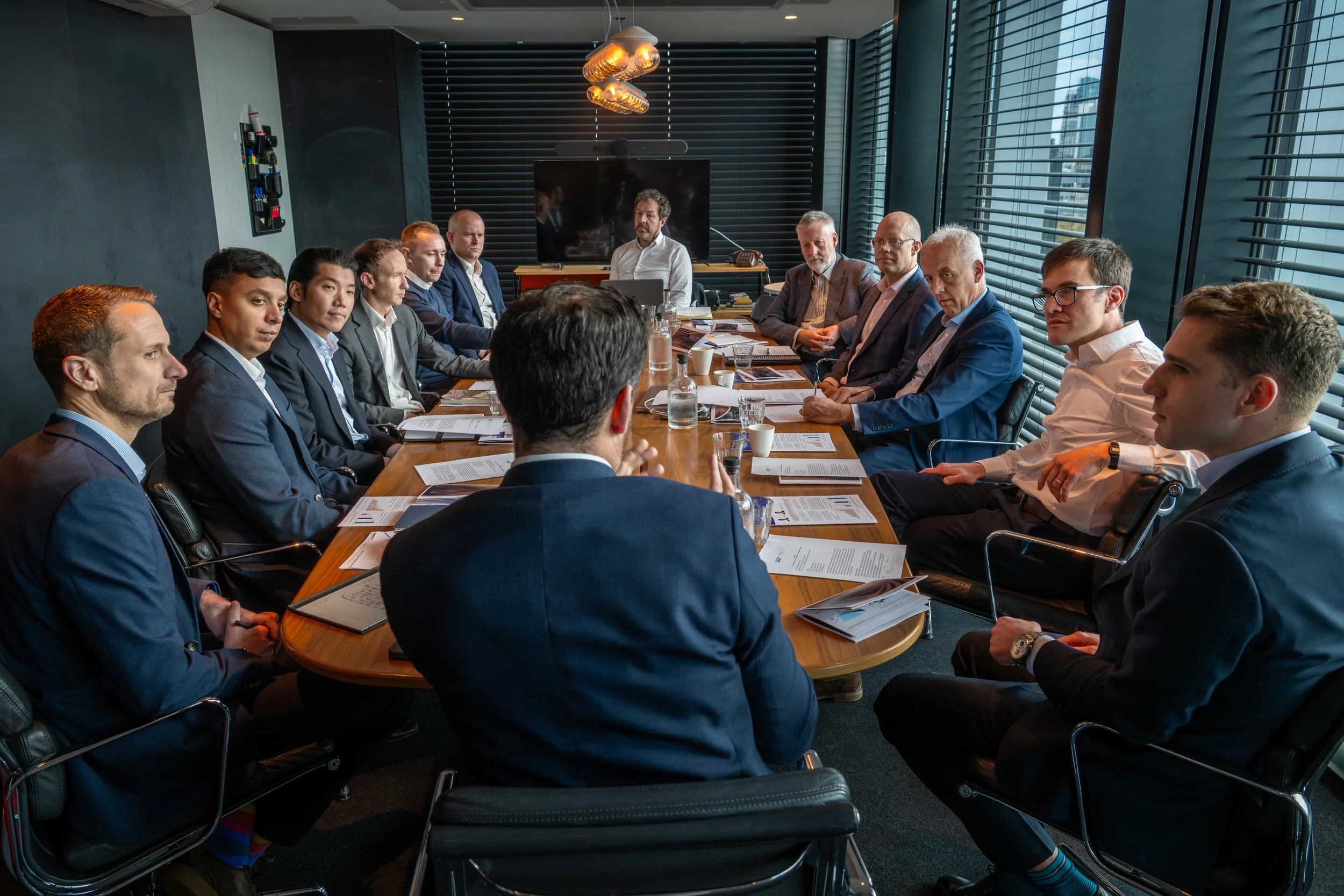 Business meeting with fifteen people seated around a large conference table in a modern office with large windows and blackout blinds.
