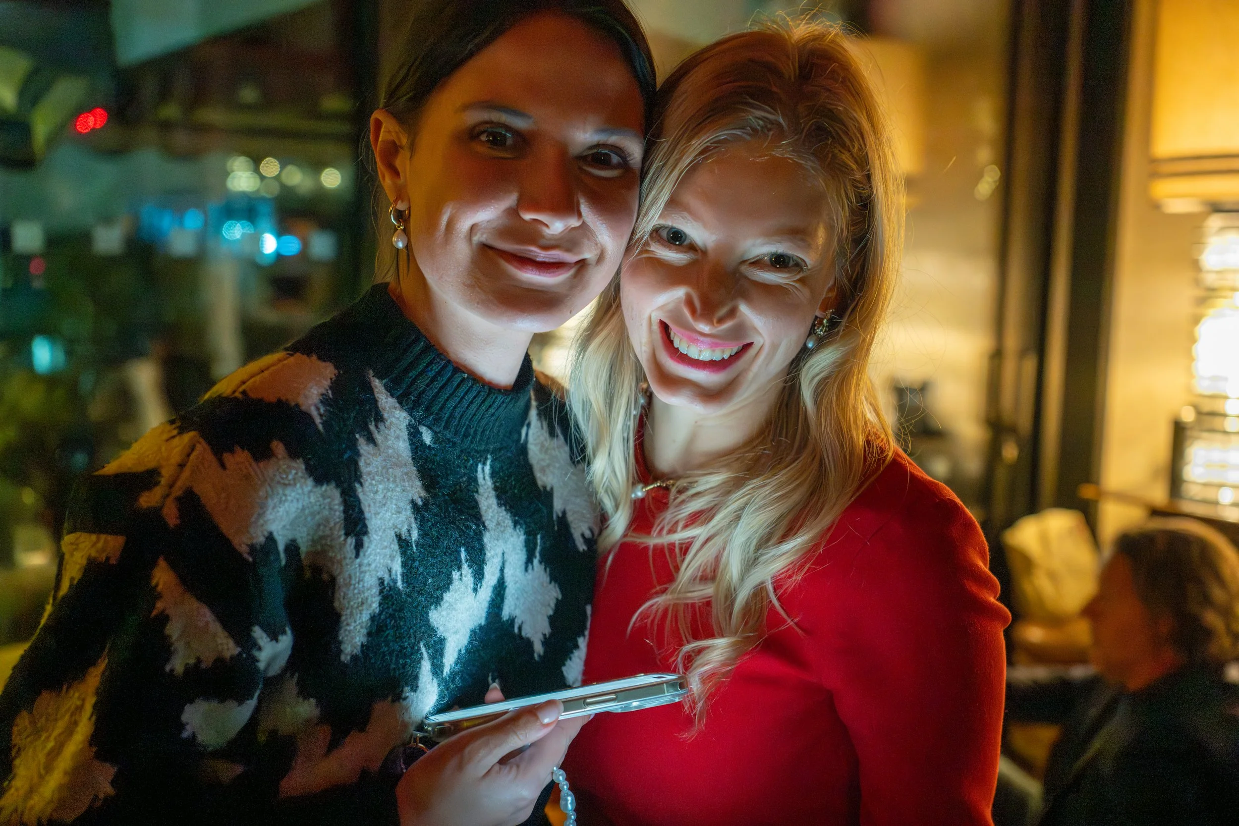 Two women in a warmly lit indoor setting smiling at the camera, one holding a smartphone, with other people blurred in the background.